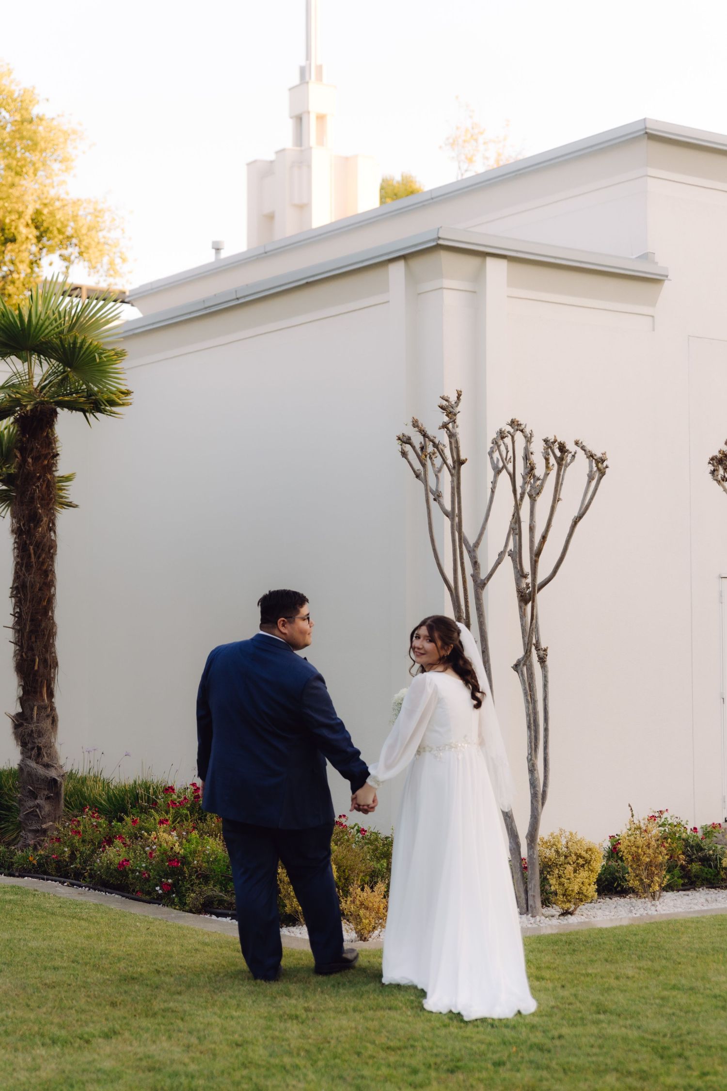 Wedding couple holds hands walking near white LDS temple with palm tree and spire visible.