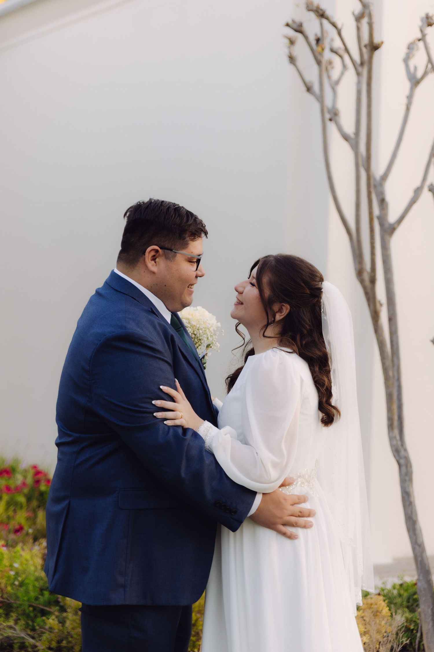 Bride in white gown and groom in navy suit embrace closely outside white temple wall.