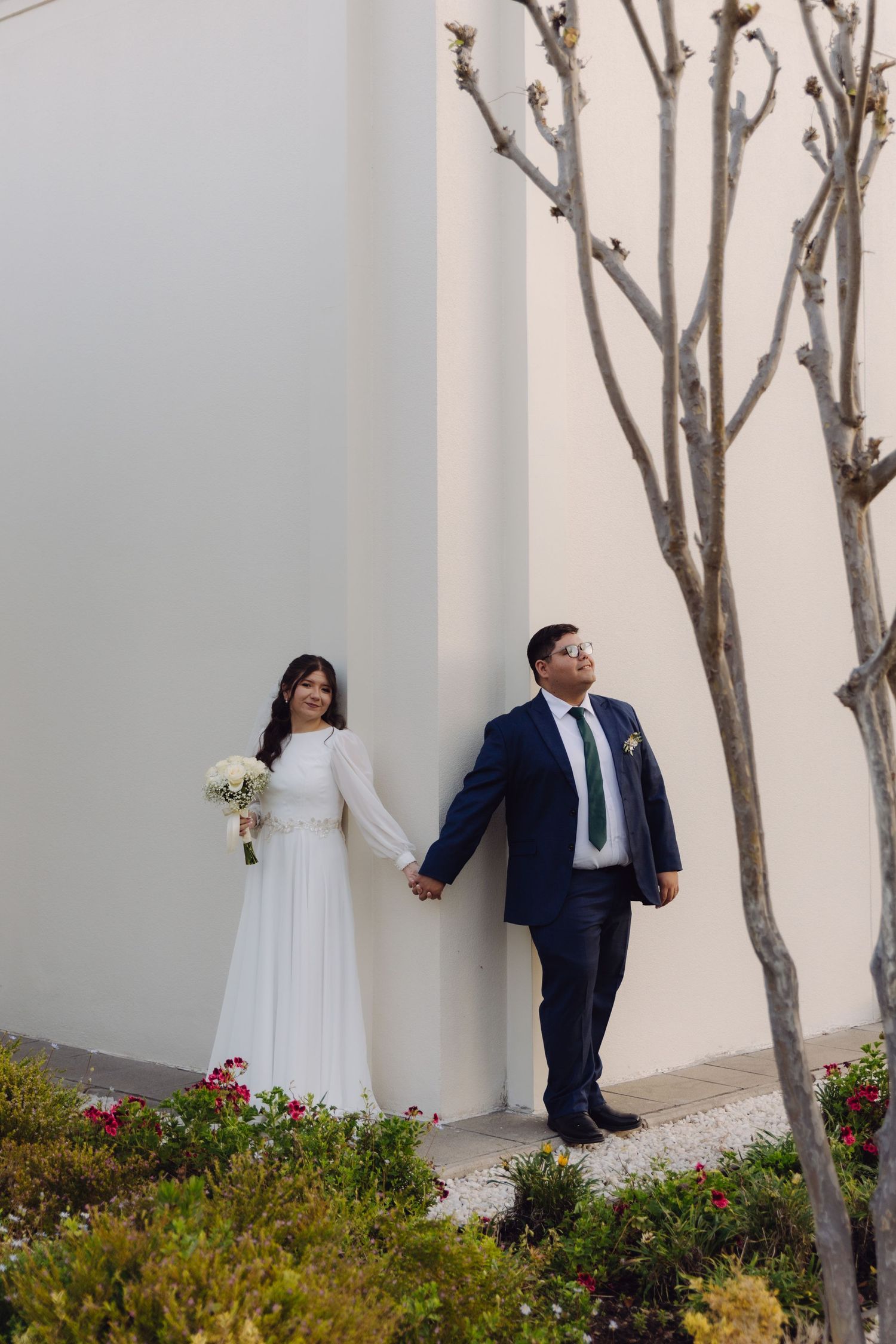 Bride holding bouquet and groom in navy suit hold hands leaning against white temple wall.