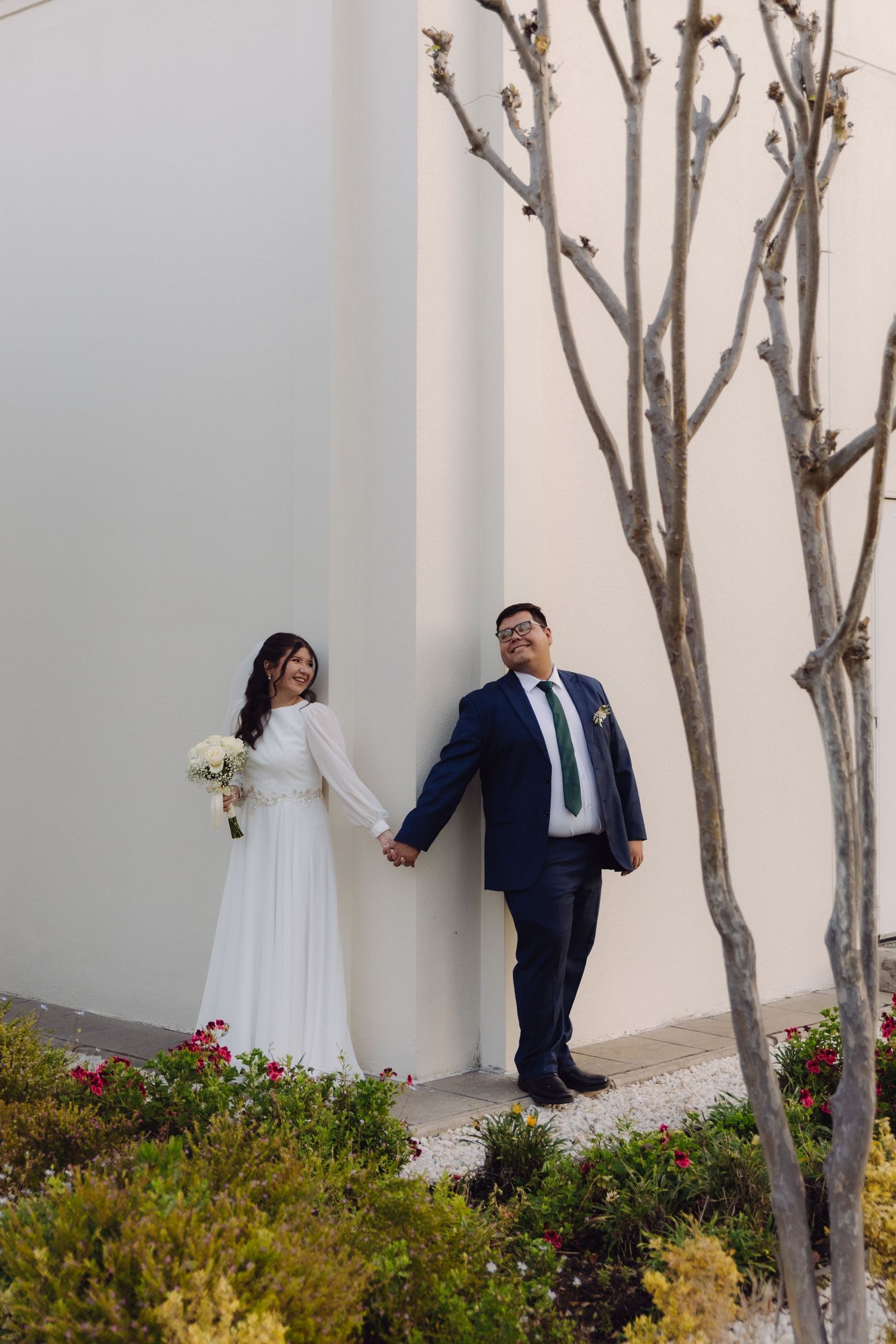 Smiling newlyweds hold hands beside bare trees against white temple exterior wall with flowers.
