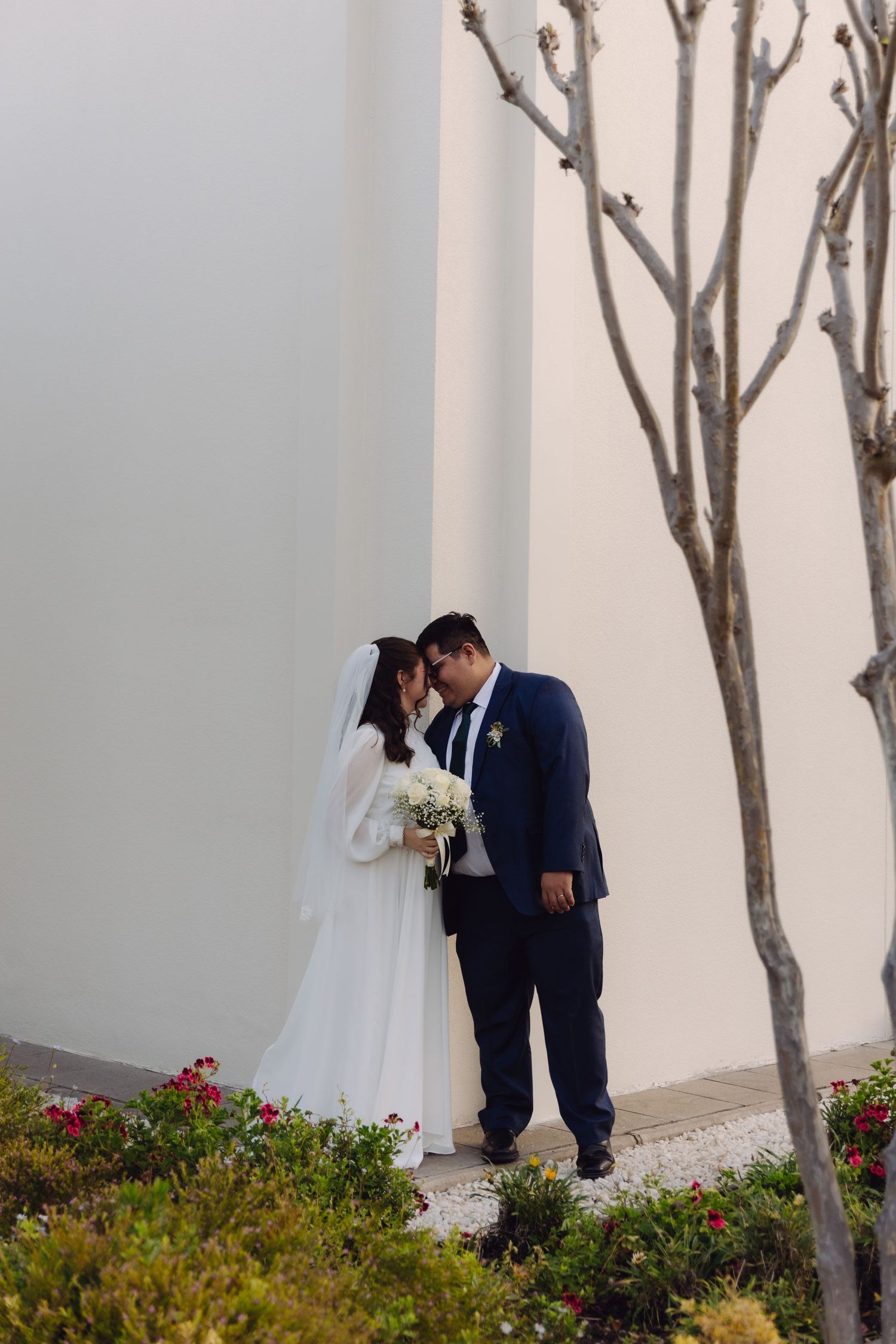 Bride and groom touch foreheads tenderly beside bare tree and white temple wall with flowers.