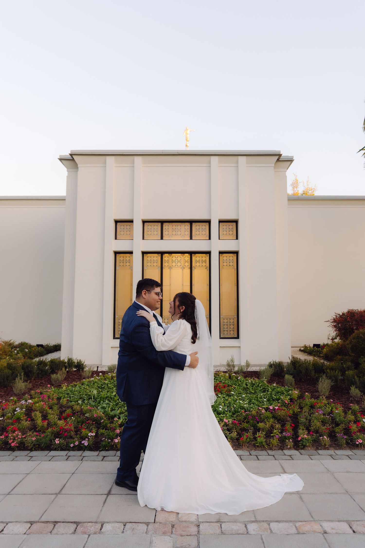 Newlyweds embrace romantically in front of white LDS temple entrance with golden windows at dusk.