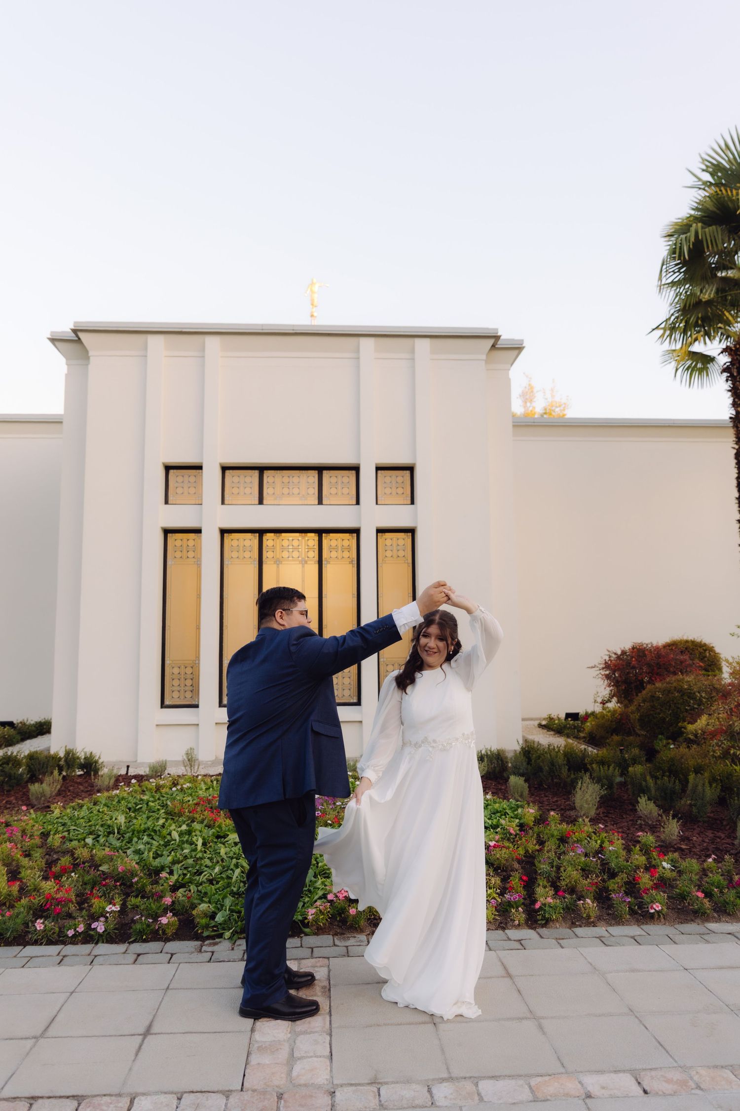 Groom twirls bride in white gown outside illuminated LDS temple with golden stained glass windows.