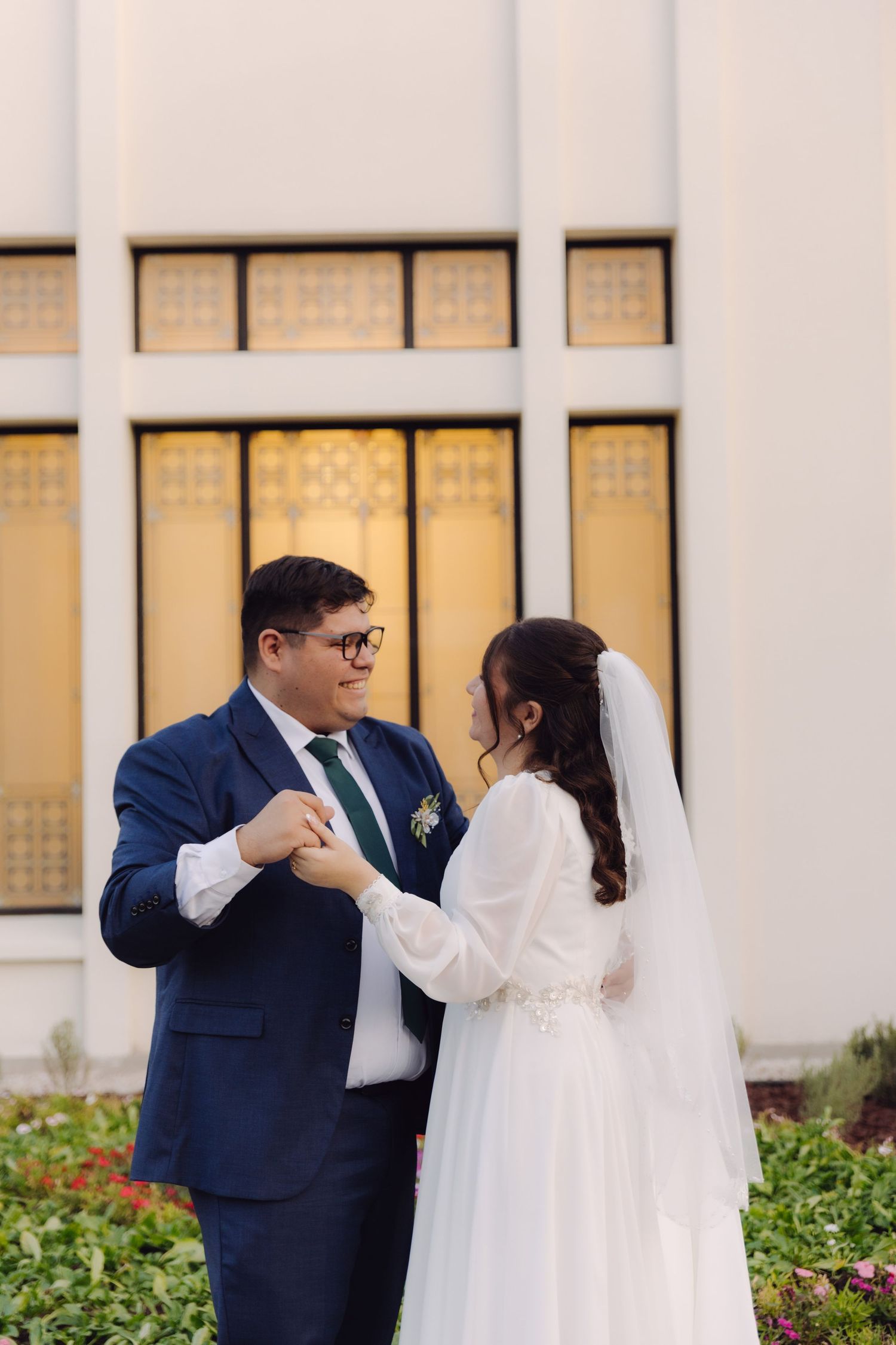 Groom in navy suit holds brides hand while gazing at her before golden temple windows.