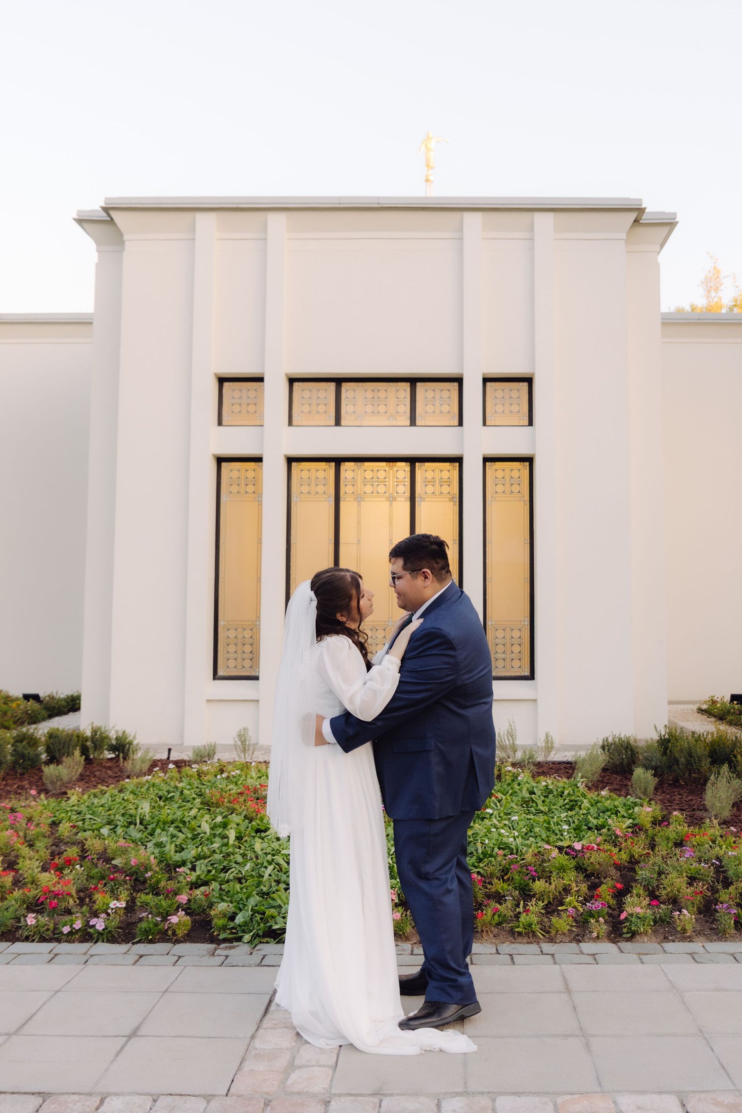 Bride and groom share intimate embrace before white LDS temple facade with glowing golden windows.