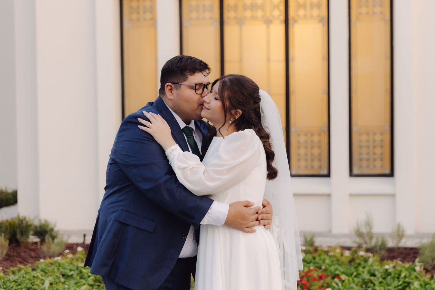 Bride and groom sharing a tender kiss outside a white temple, groom in navy suit, bride in elegant white gown with veil.