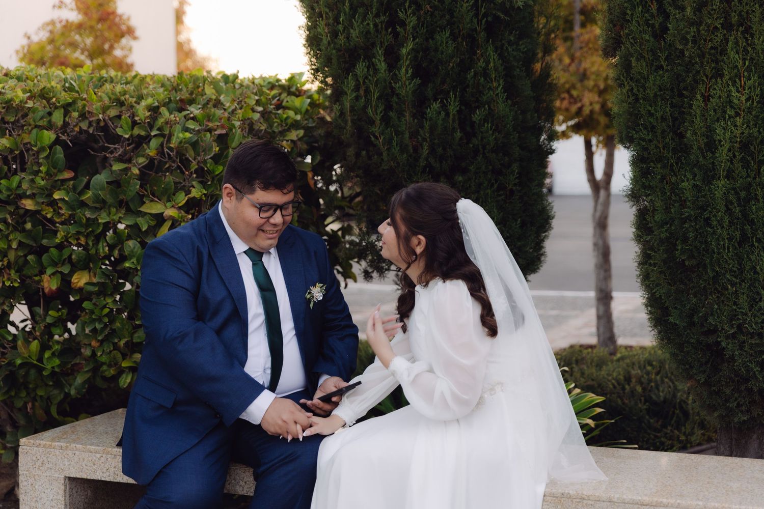 Bride and groom sitting on a stone bench in a garden, holding hands and sharing an intimate moment.