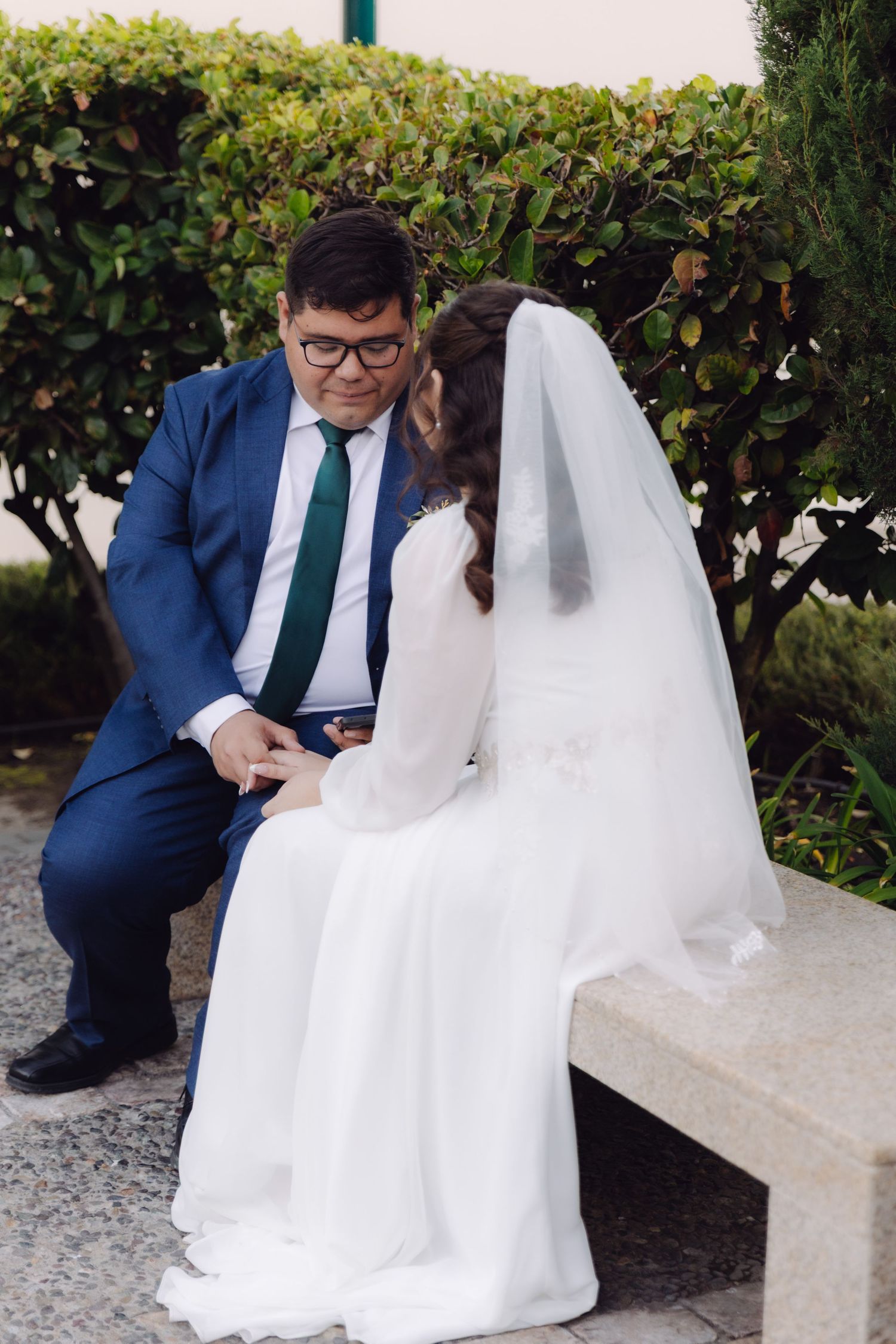 Groom in navy suit kneeling beside bride in white dress seated on stone bench surrounded by lush greenery.