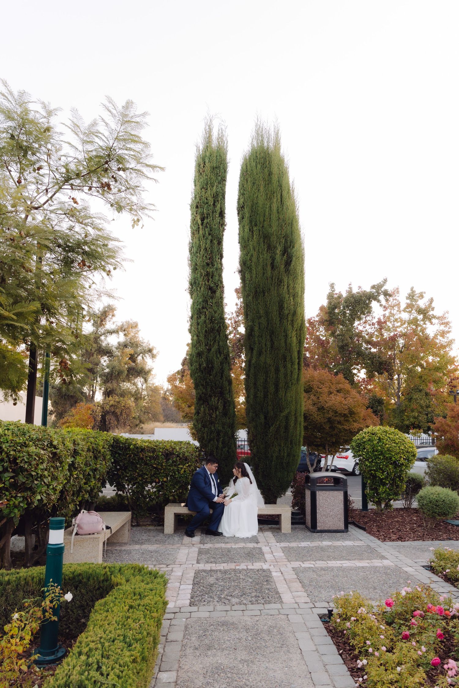 Bride and groom sitting together on a garden bench framed by tall cypress trees during autumn wedding.