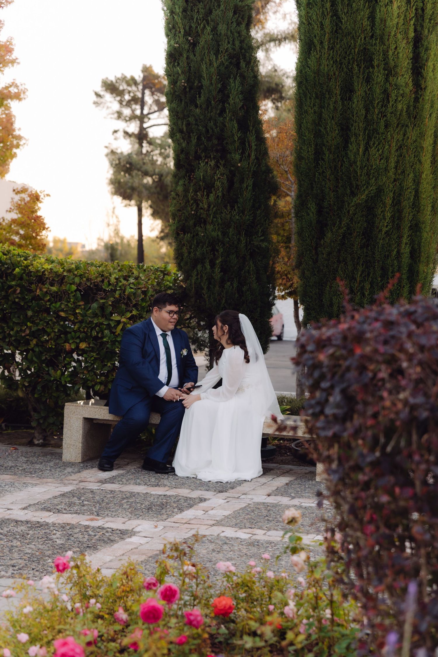 Bride and groom sharing an intimate conversation on a bench surrounded by cypress trees and colorful roses.