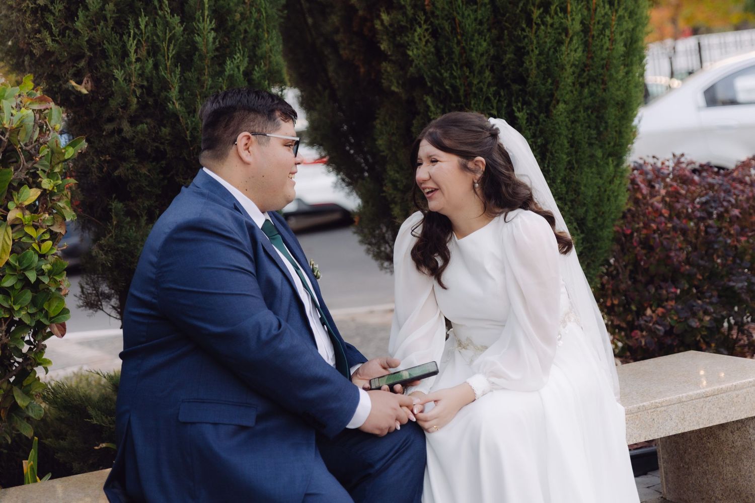 Smiling bride in white veil and groom in navy suit holding hands and laughing together on a bench.