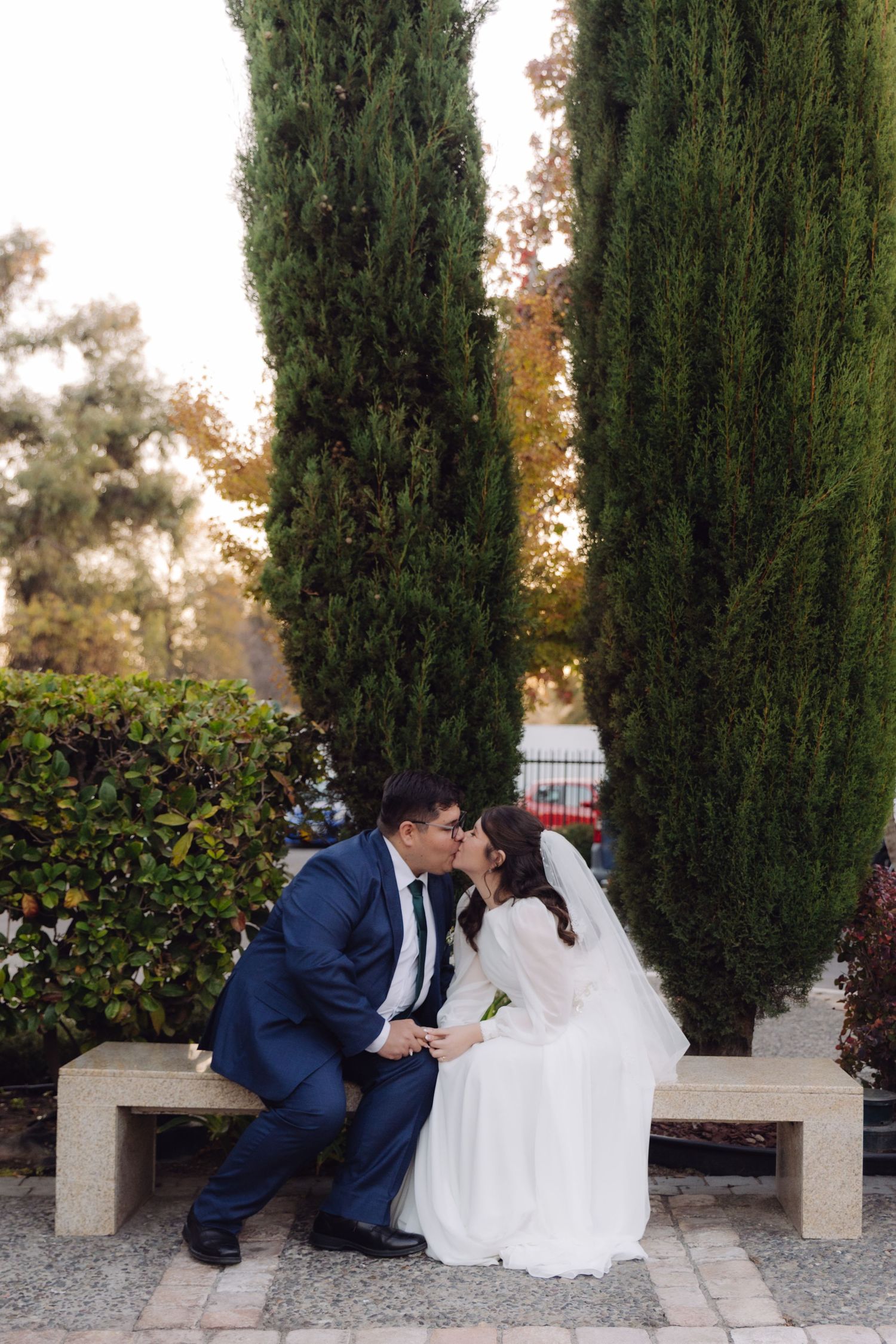 Bride and groom sharing a romantic kiss on a stone bench flanked by tall cypress trees in a garden.