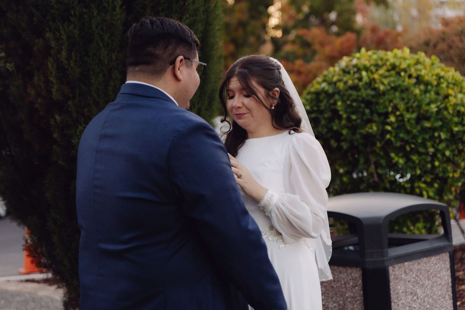 Bride smiles emotionally as groom adjusts her boutonniere in a garden setting.