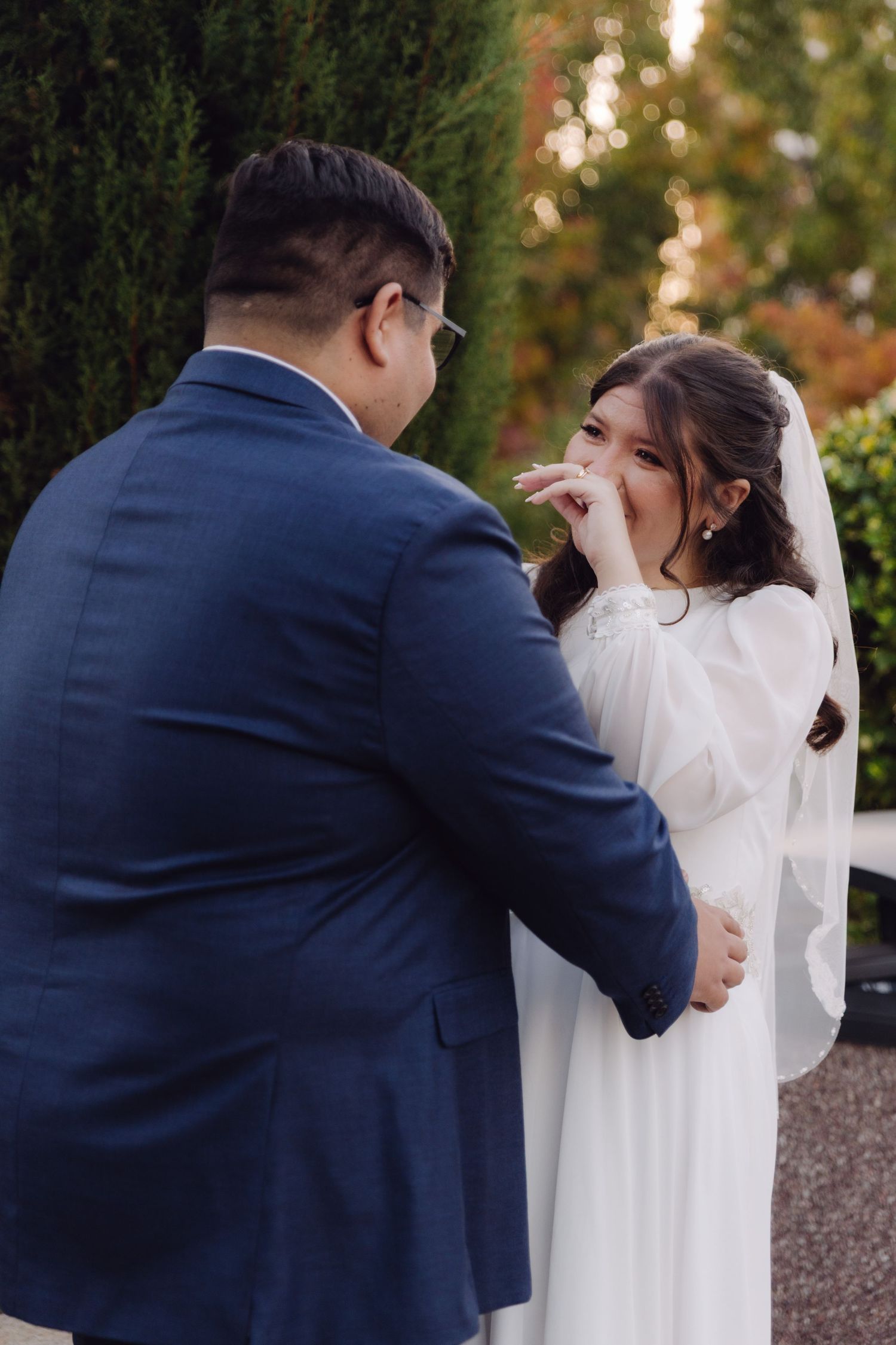 Emotional bride covers mouth while groom holds her hands during outdoor wedding moment.