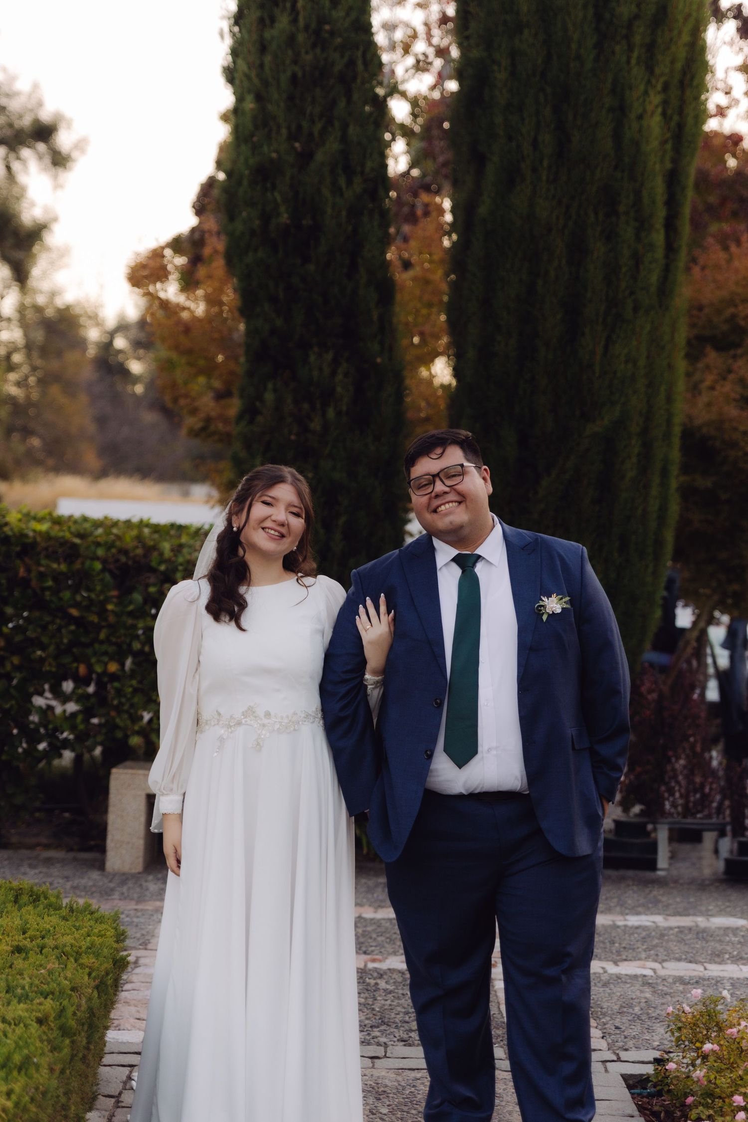Smiling bride and groom walk together in garden with tall cypress trees backdrop.