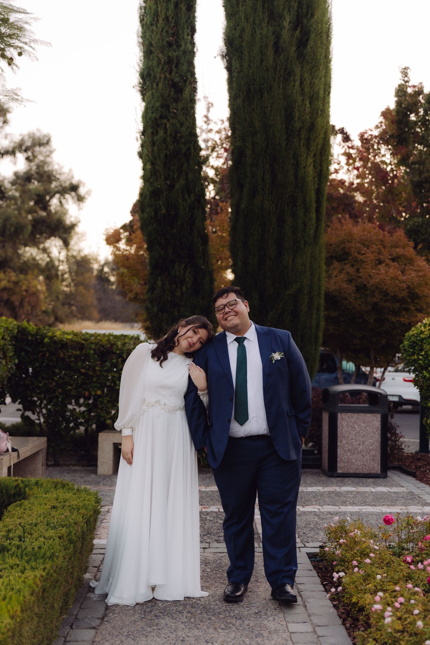 Bride leans on grooms shoulder as they pose on garden path at sunset.
