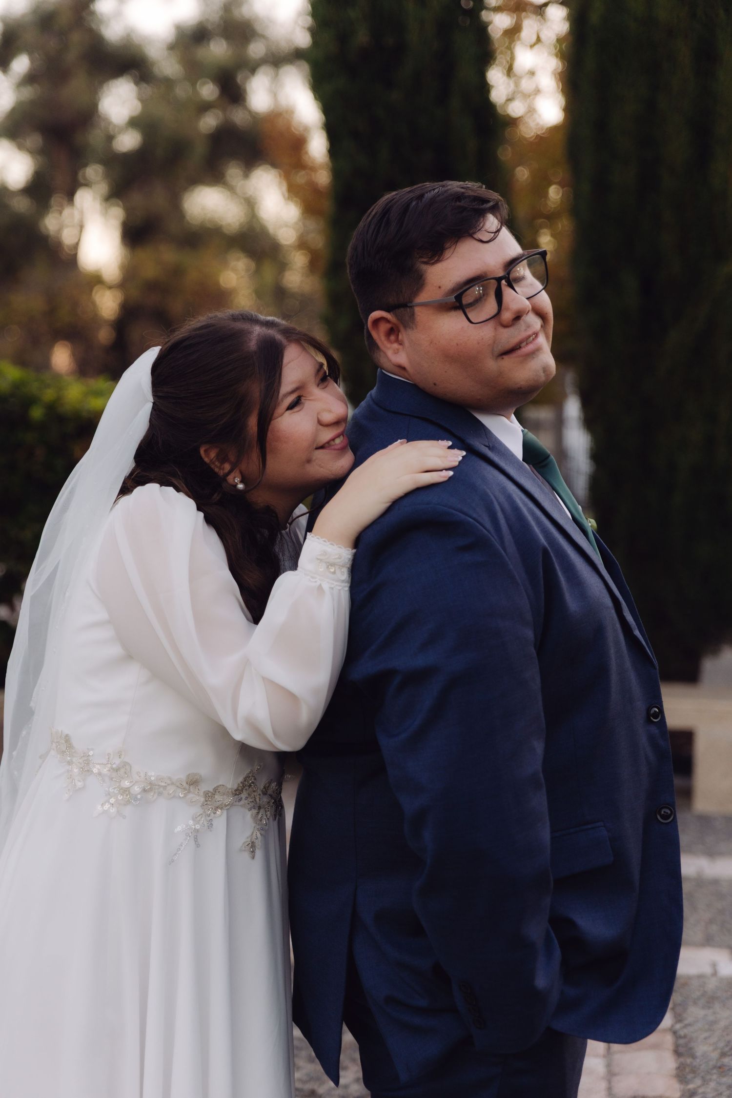 Bride embraces groom from behind as he smiles in a garden wedding portrait.