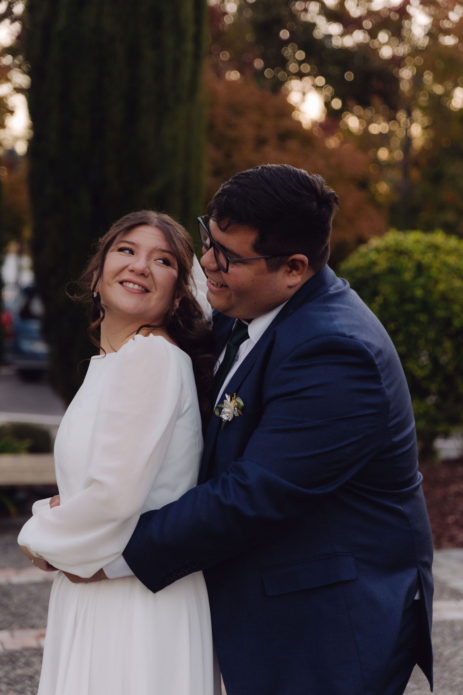 Groom whispers to laughing bride while embracing her in an outdoor wedding portrait.