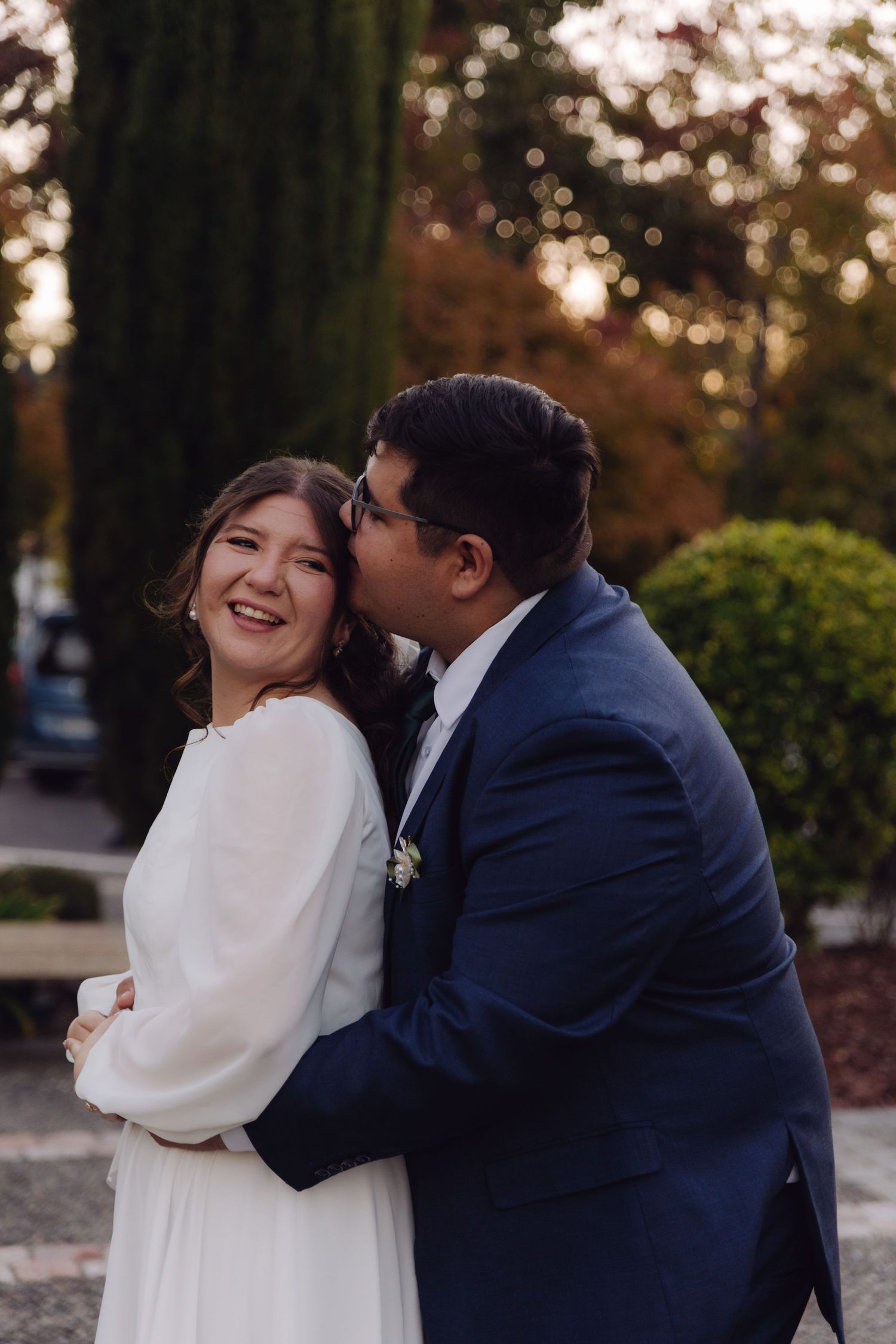 Bride in white dress laughing joyfully as groom in navy suit kisses her cheek outdoors.