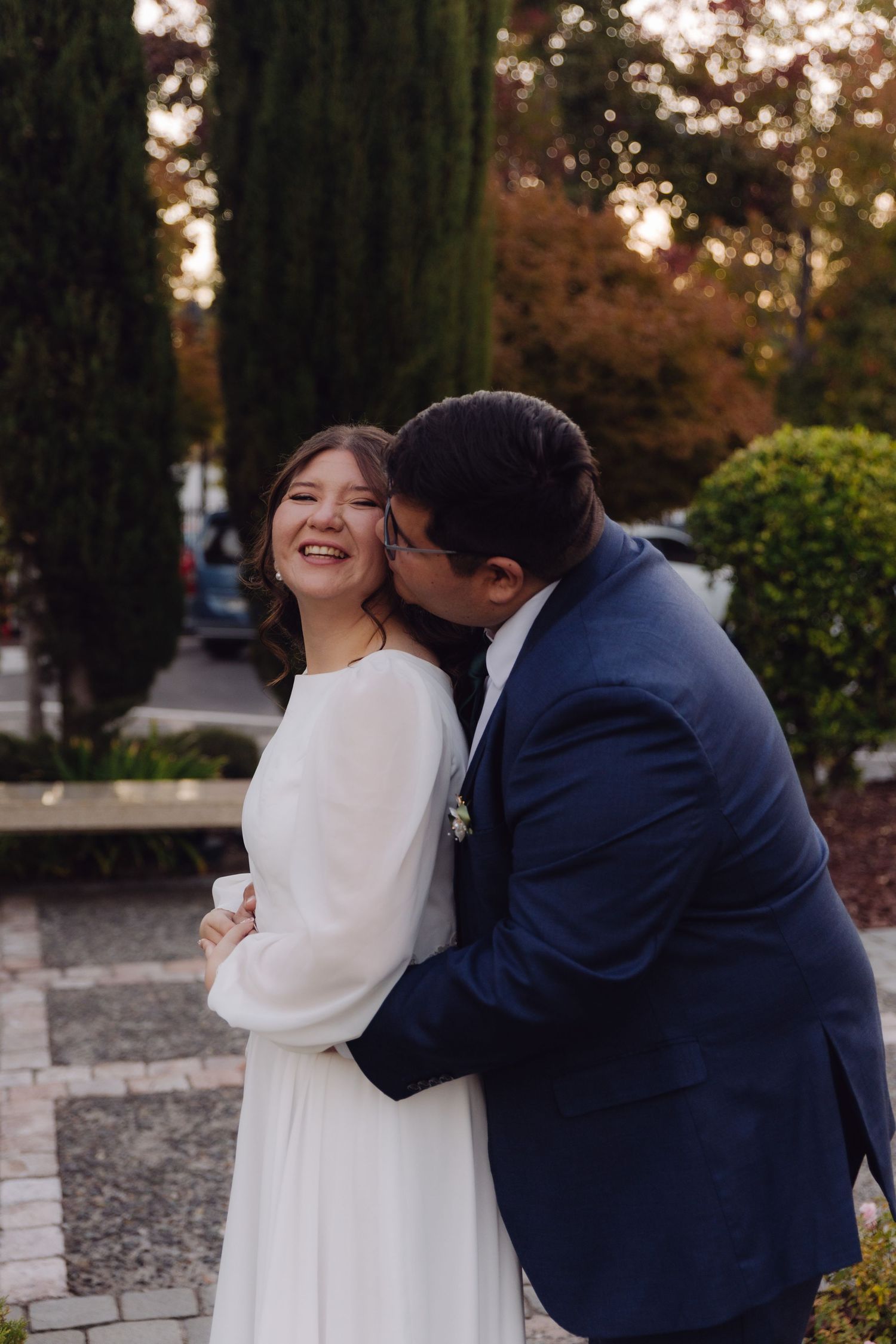 Happy bride in long white dress smiling as groom in navy suit embraces and kisses her cheek.