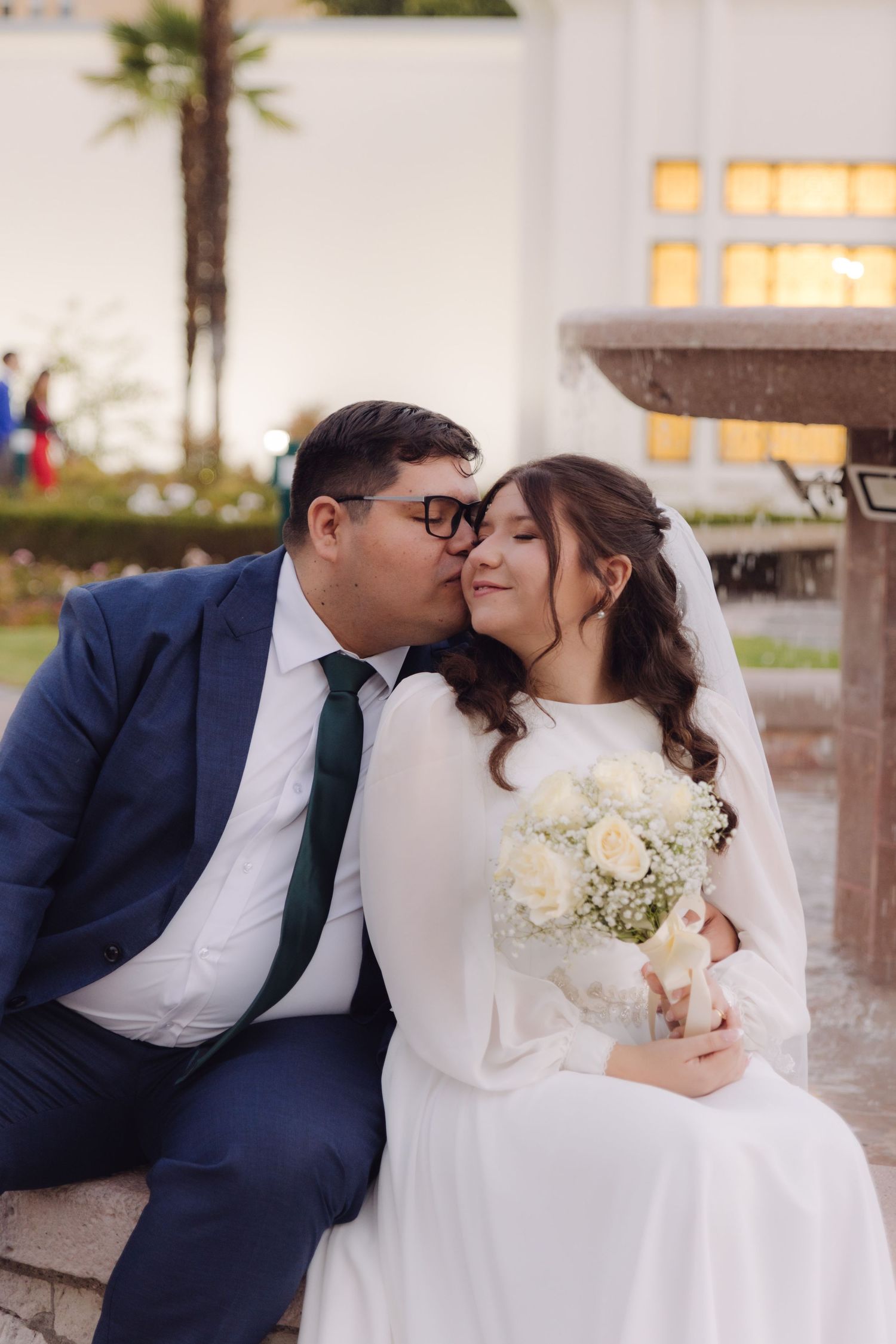 Groom in navy suit kissing bride on cheek as she holds white rose bouquet by a fountain.