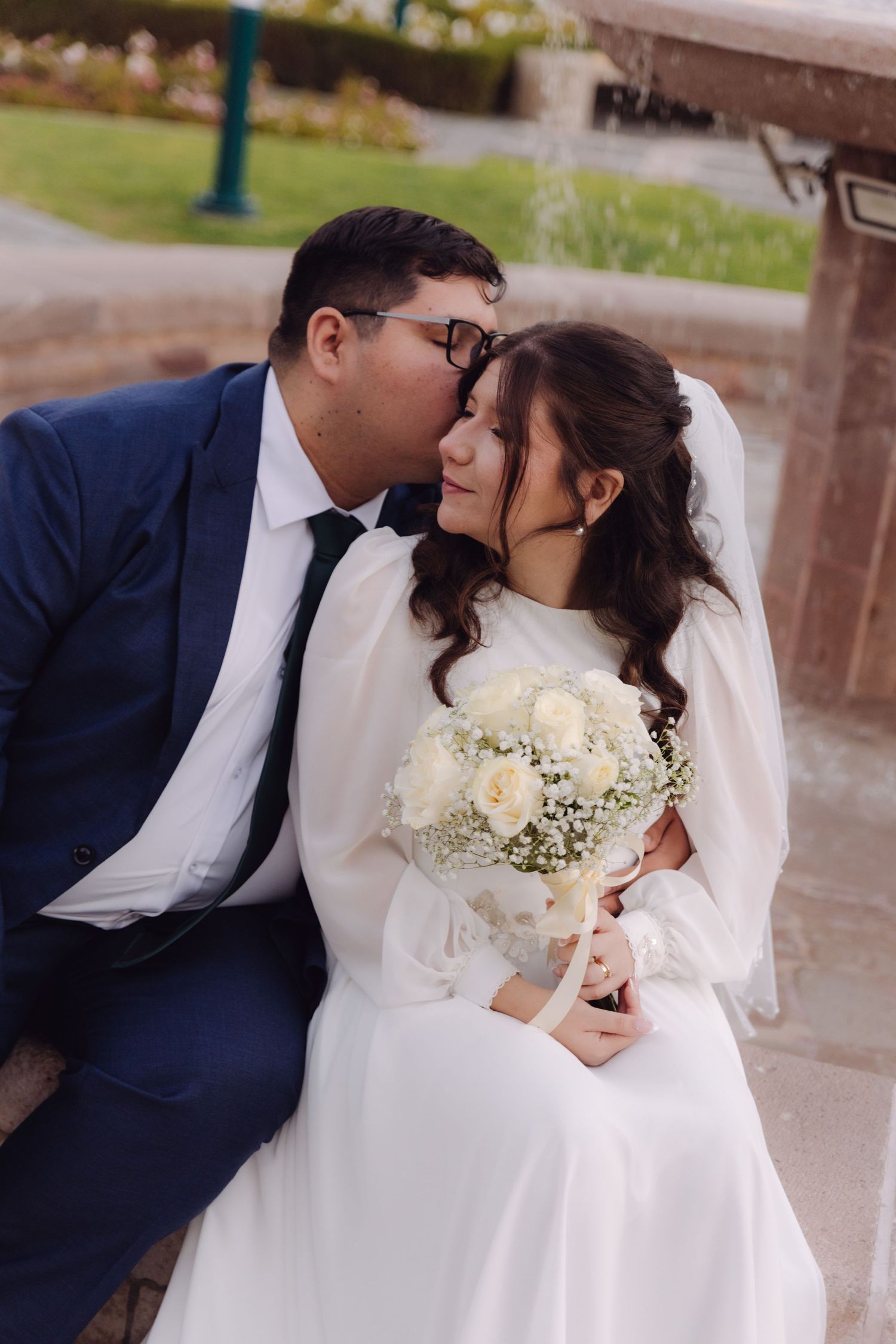 Bride and groom sharing a tender kiss near a fountain, bride holding a white rose bouquet.