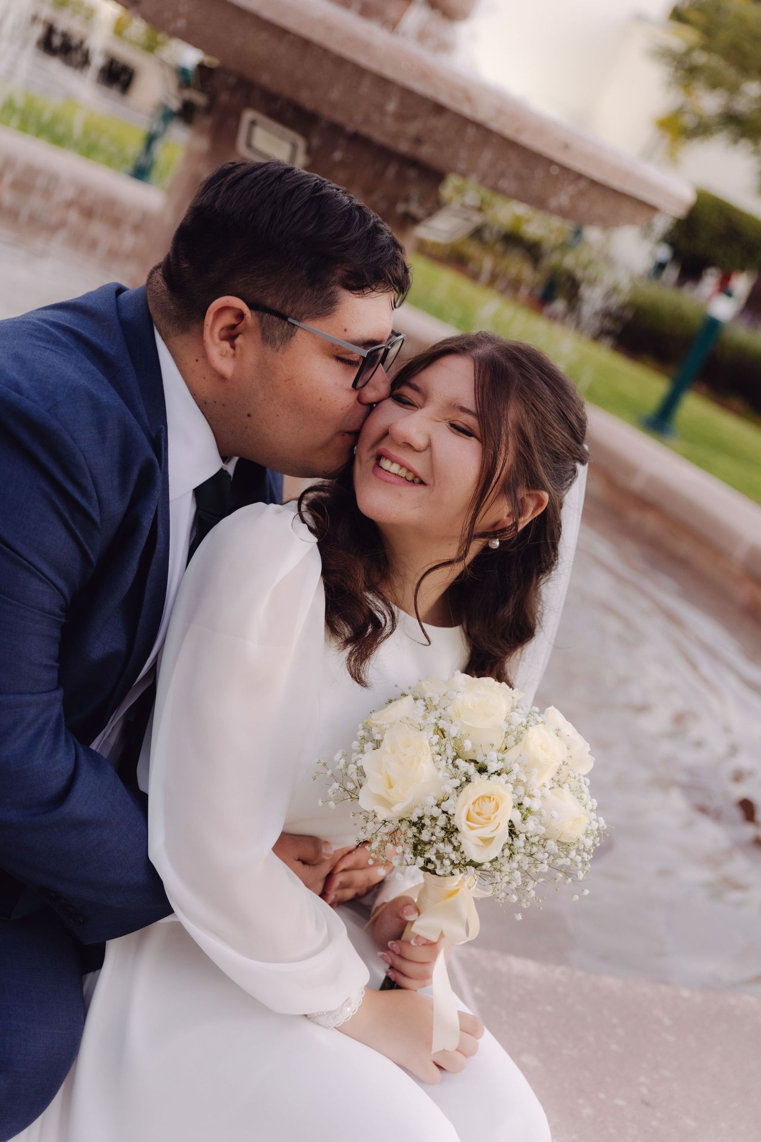 Groom kissing smiling bride on the cheek near a fountain, holding white roses.