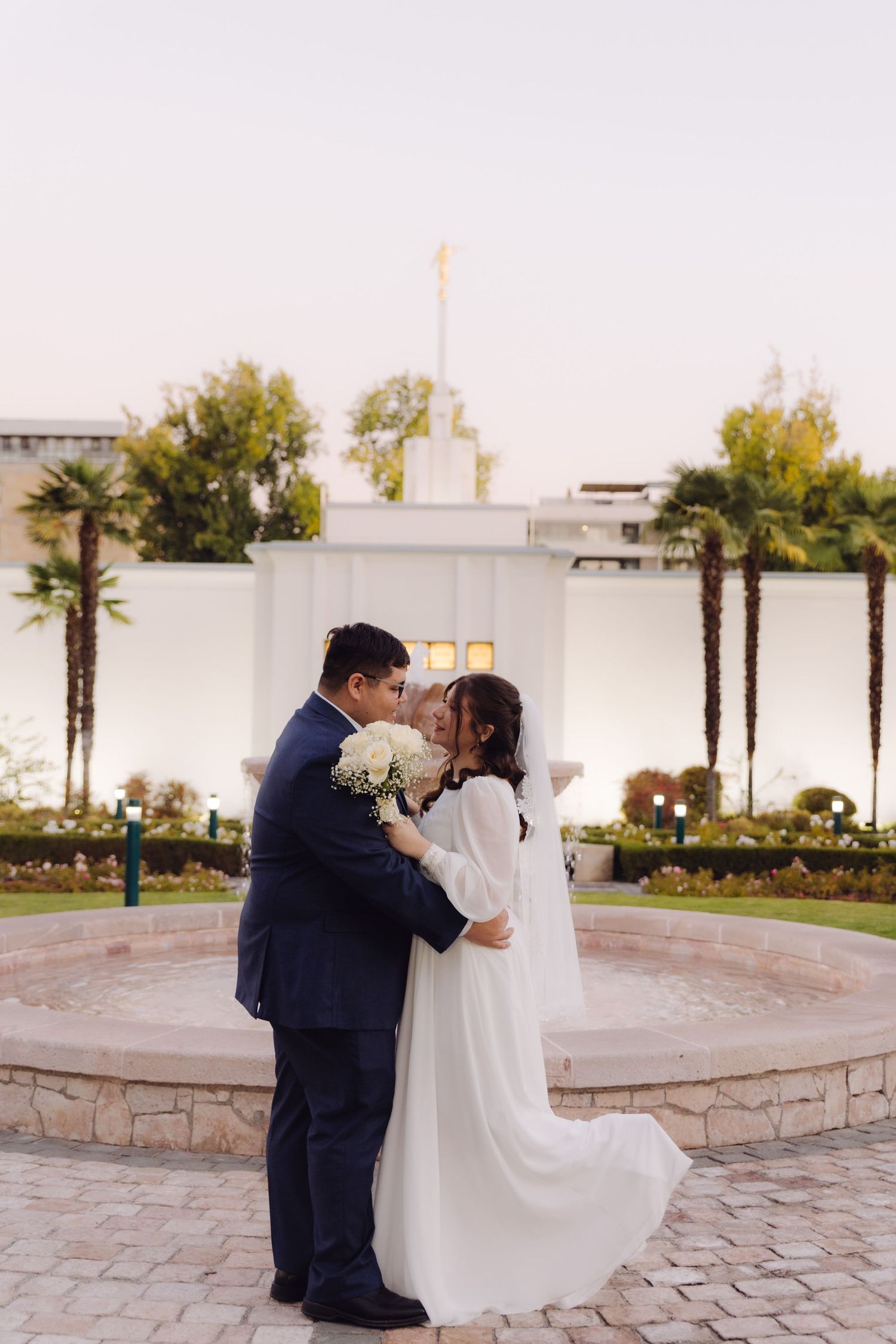 Bride and groom embracing near a fountain with an LDS temple in the background.