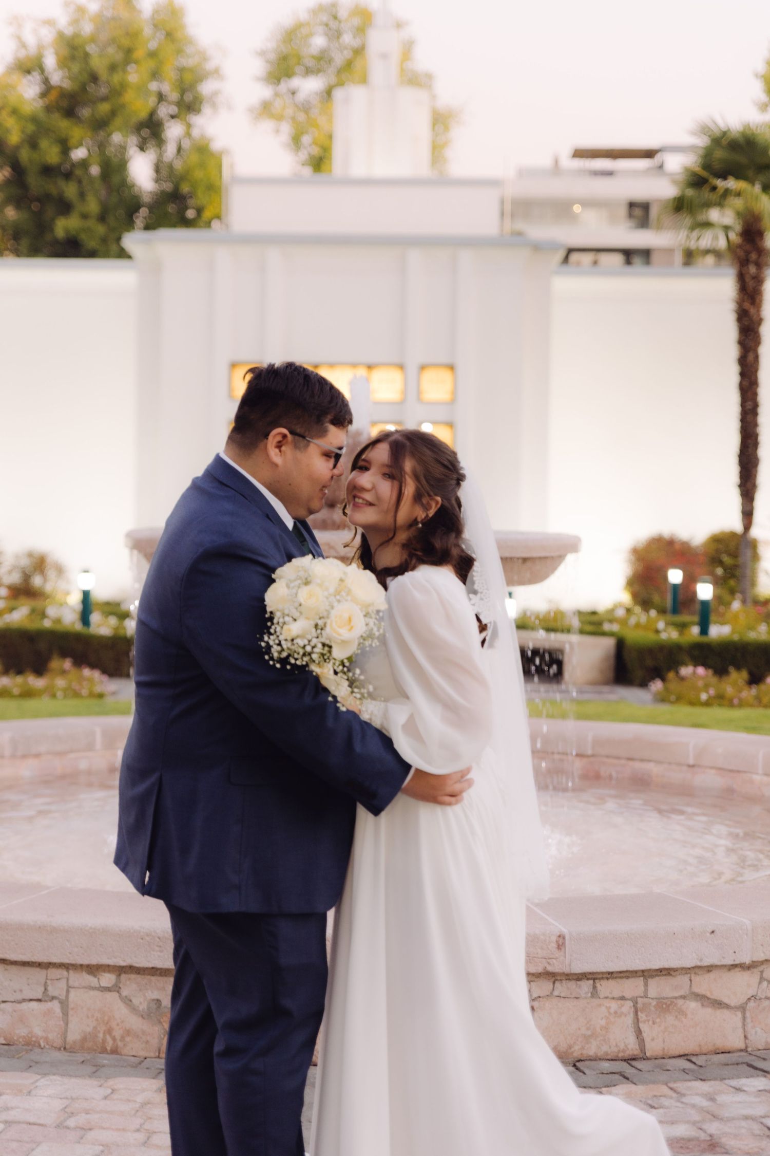 Couple sharing a tender embrace near a stone fountain outside an LDS temple.