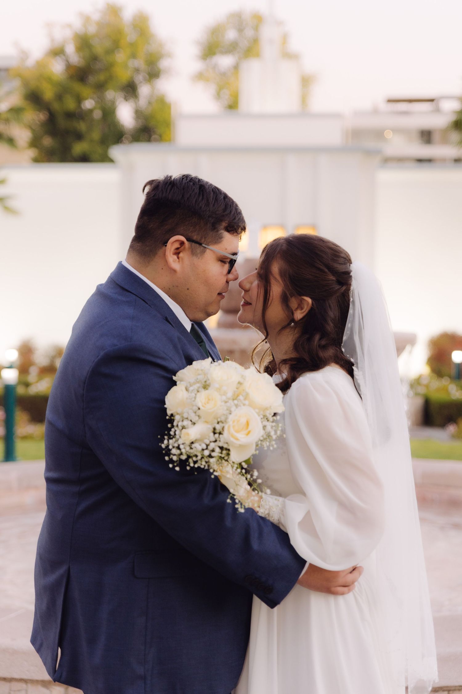 Bride and groom leaning in for a kiss outdoors, holding a white rose bouquet.