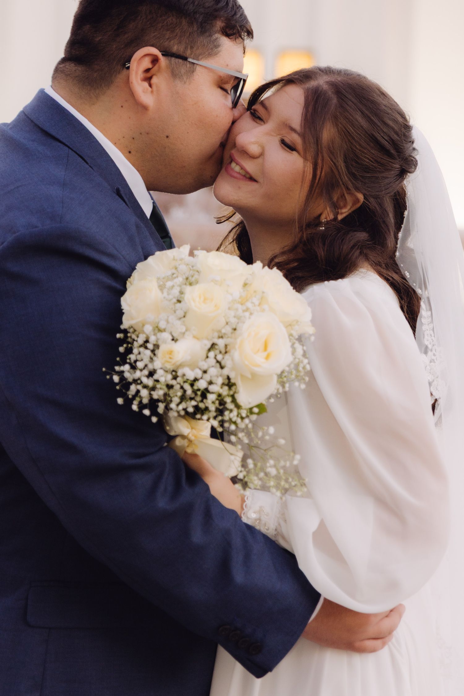 Groom in navy suit kissing bride's cheek as she smiles holding white rose bouquet with gypsophila.