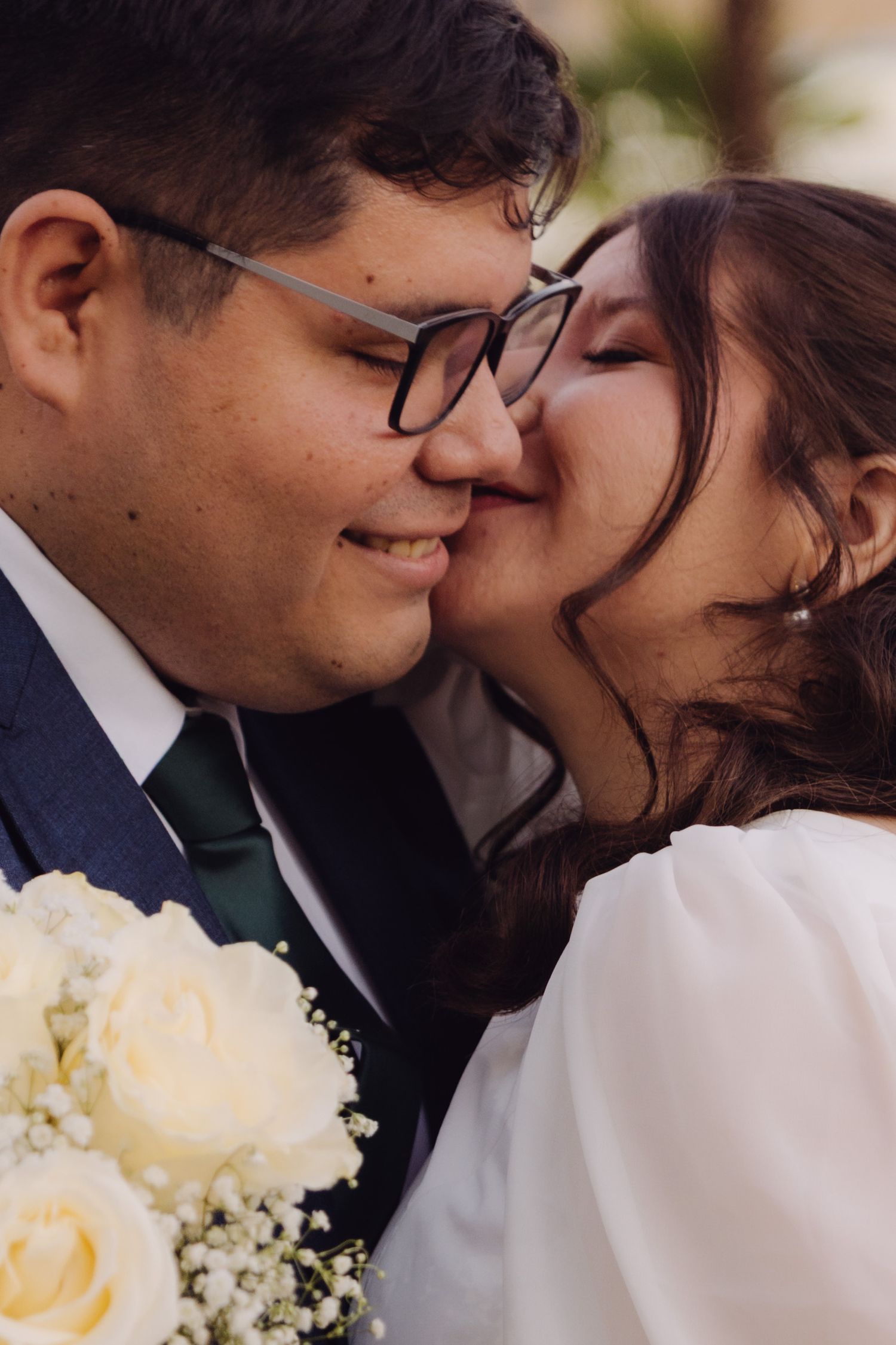 Close-up of wedding couple sharing a tender kiss, groom wearing glasses holding white rose bouquet.
