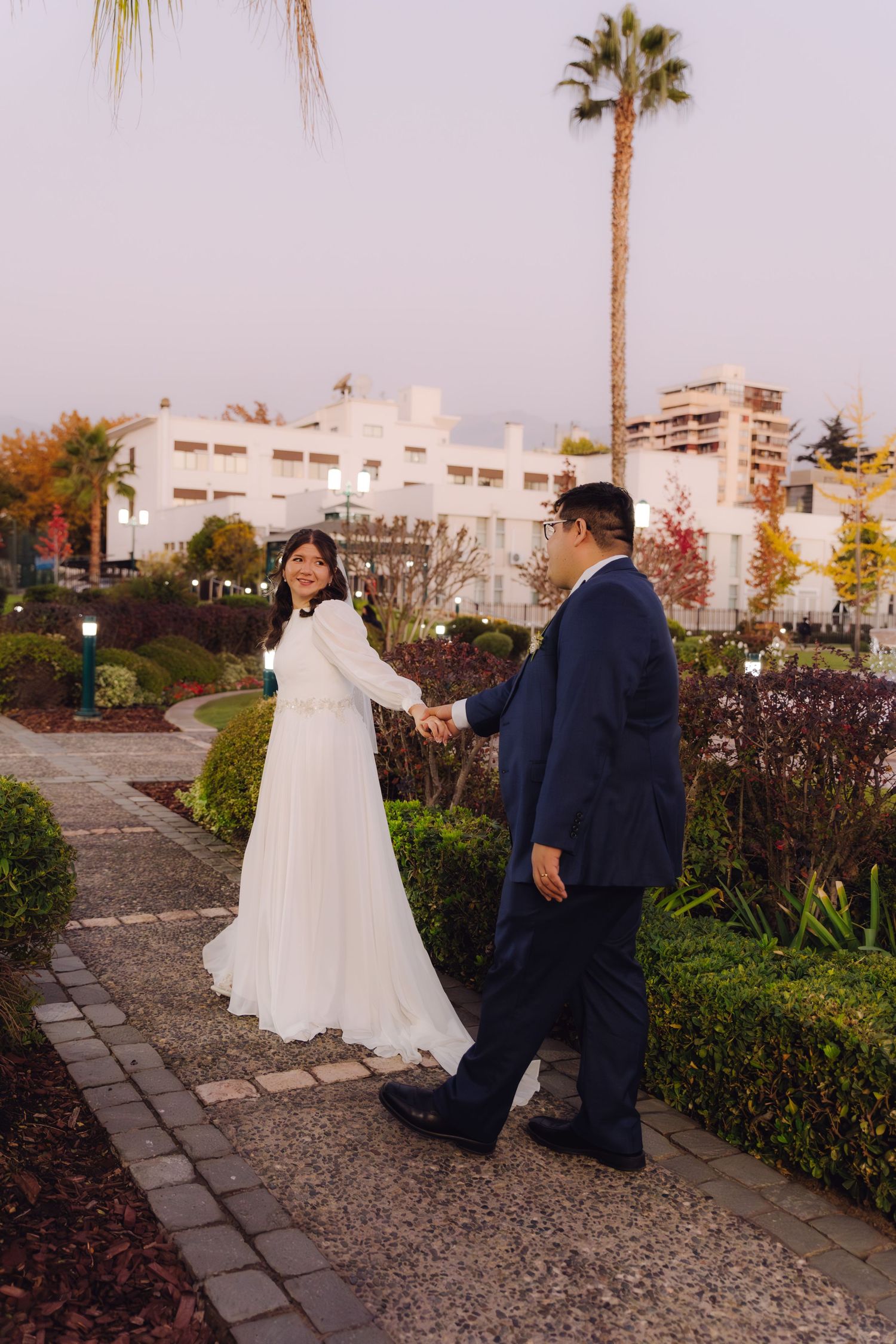 Bride and groom holding hands walking along garden path at dusk with palm trees and white building.