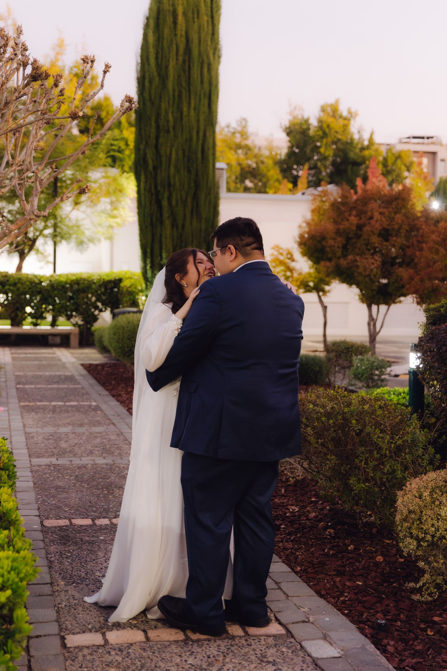 Bride and groom sharing an intimate embrace on a garden pathway surrounded by lush greenery at dusk.