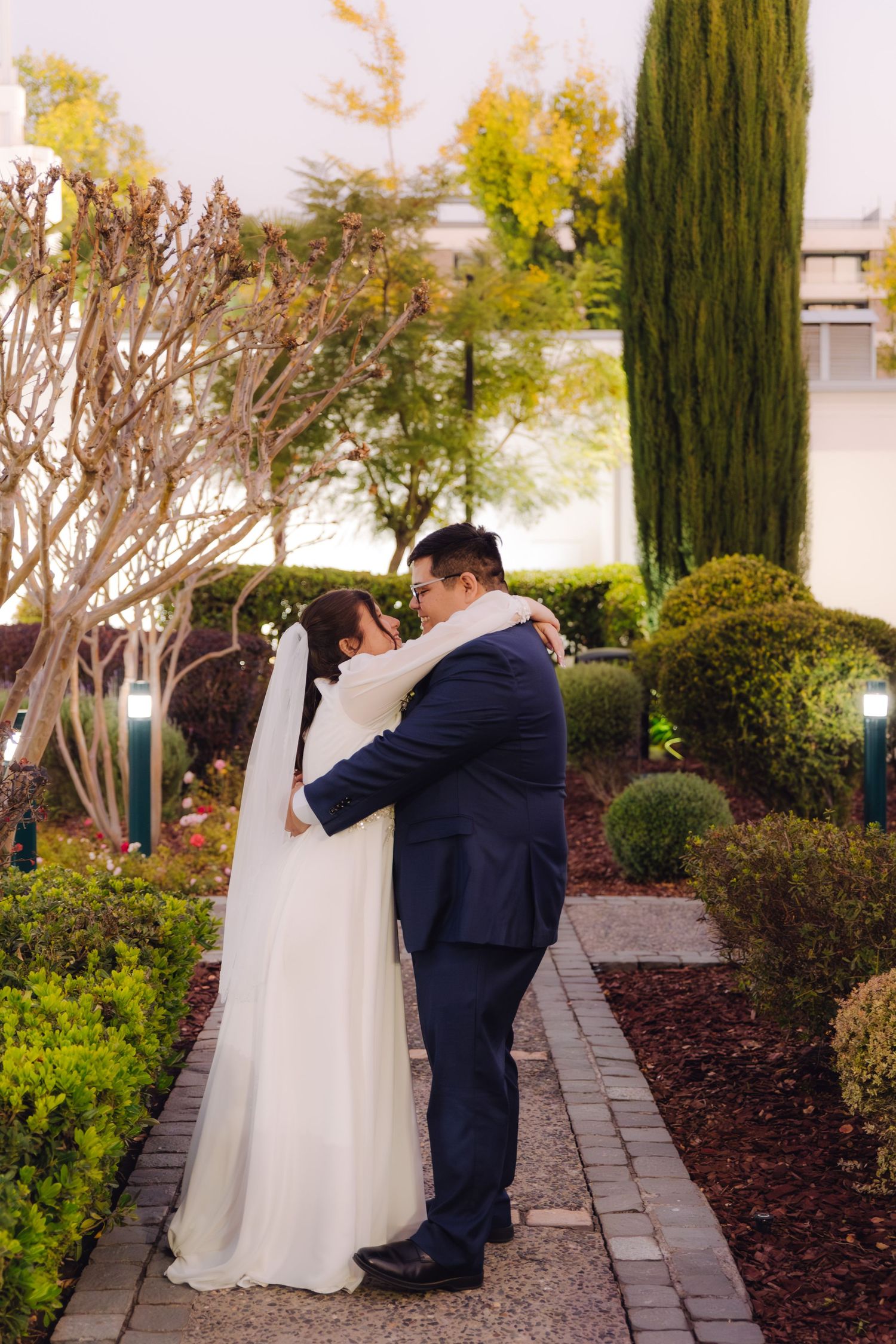 Newlywed couple embracing on a stone garden path surrounded by manicured hedges and cypress trees.