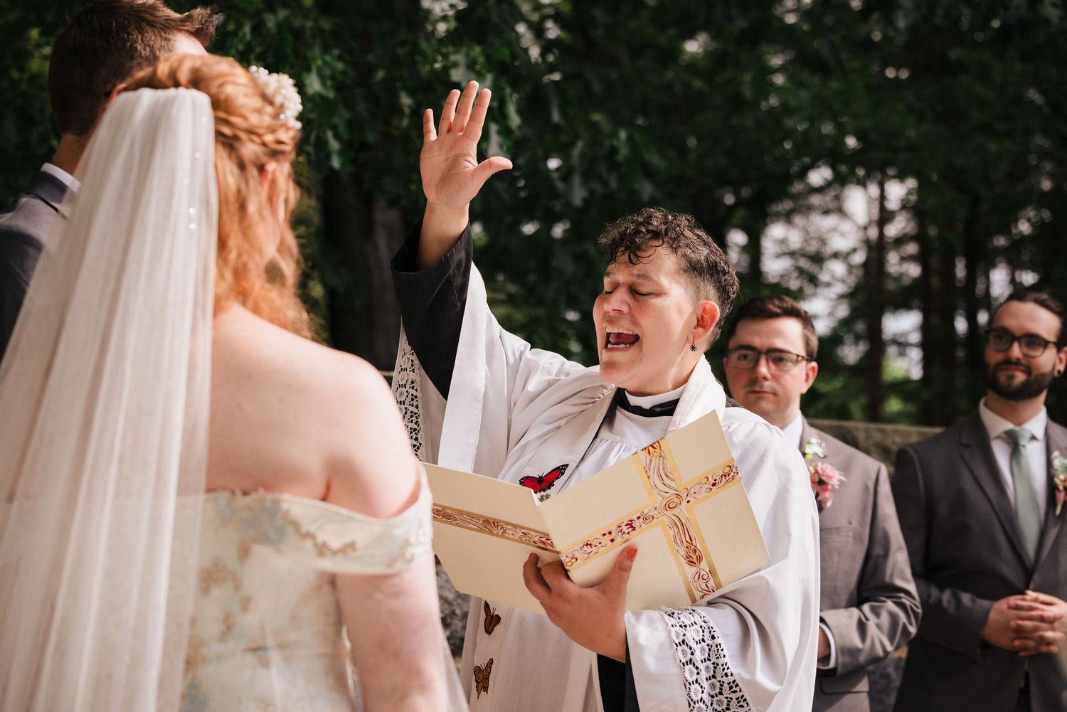 Officiant raising her hand and laughing during a joyful blessing, bride and groom standing before her.