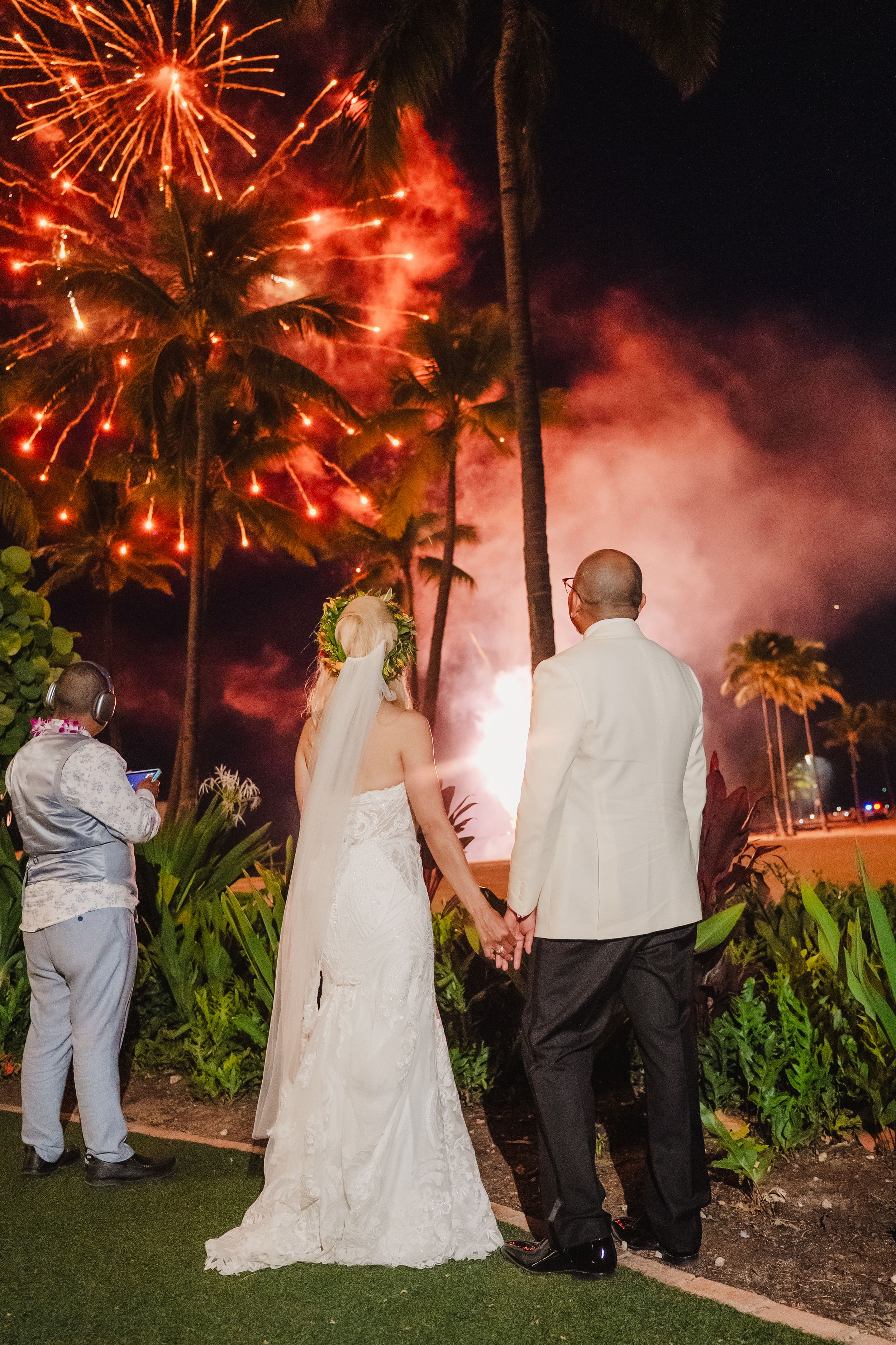 Bride and groom hold hands watching vibrant fireworks display at tropical Hawaii wedding celebration at night.