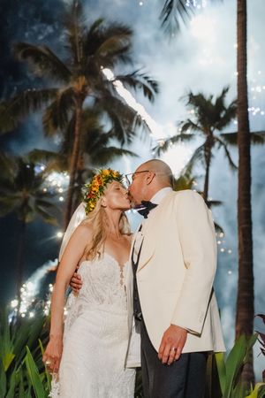 Bride and groom share a kiss under fireworks and palm trees, bride wearing floral crown and lace gown.