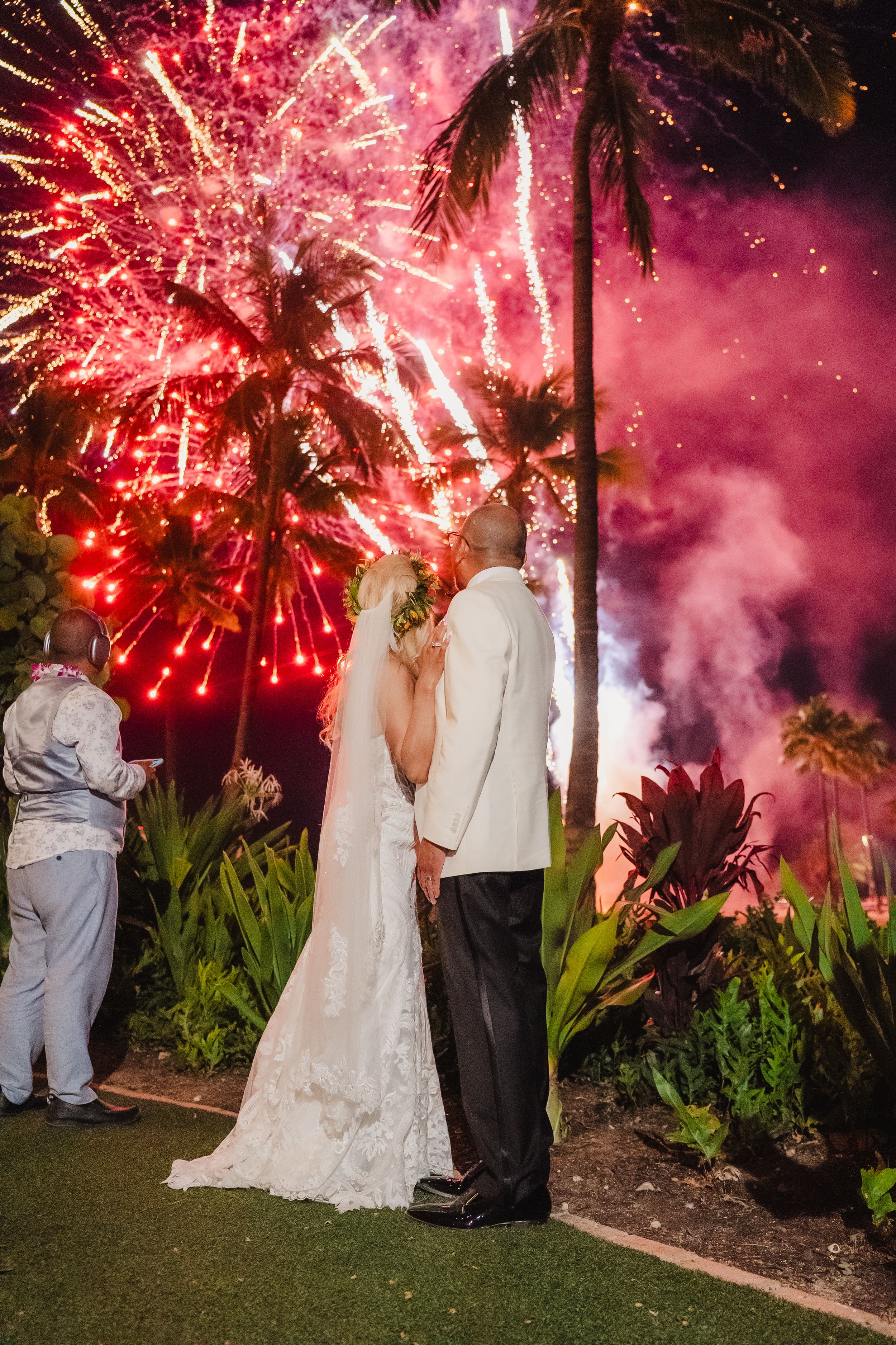 Bride and groom watch stunning red fireworks display at tropical wedding venue with palm trees at night.