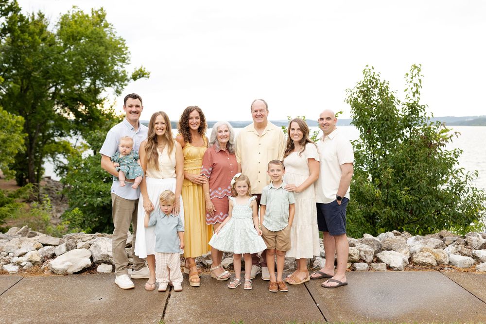 Large family group portrait smiling outdoors near a lake with lush green trees in the background on a sunny day.