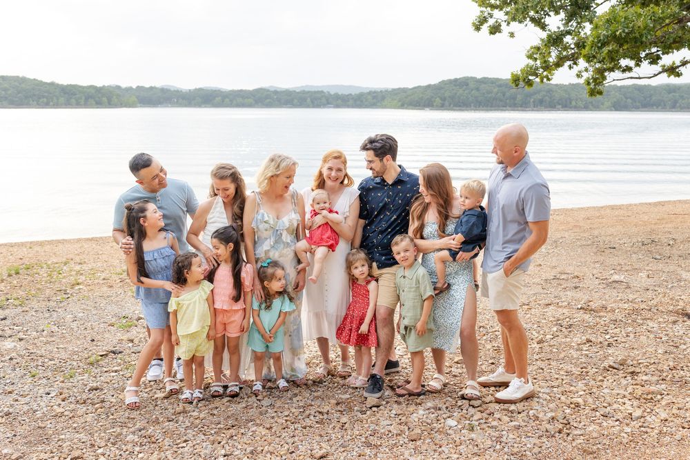 Large family group portrait with children and adults posing on a lakeside beach surrounded by lush green trees.