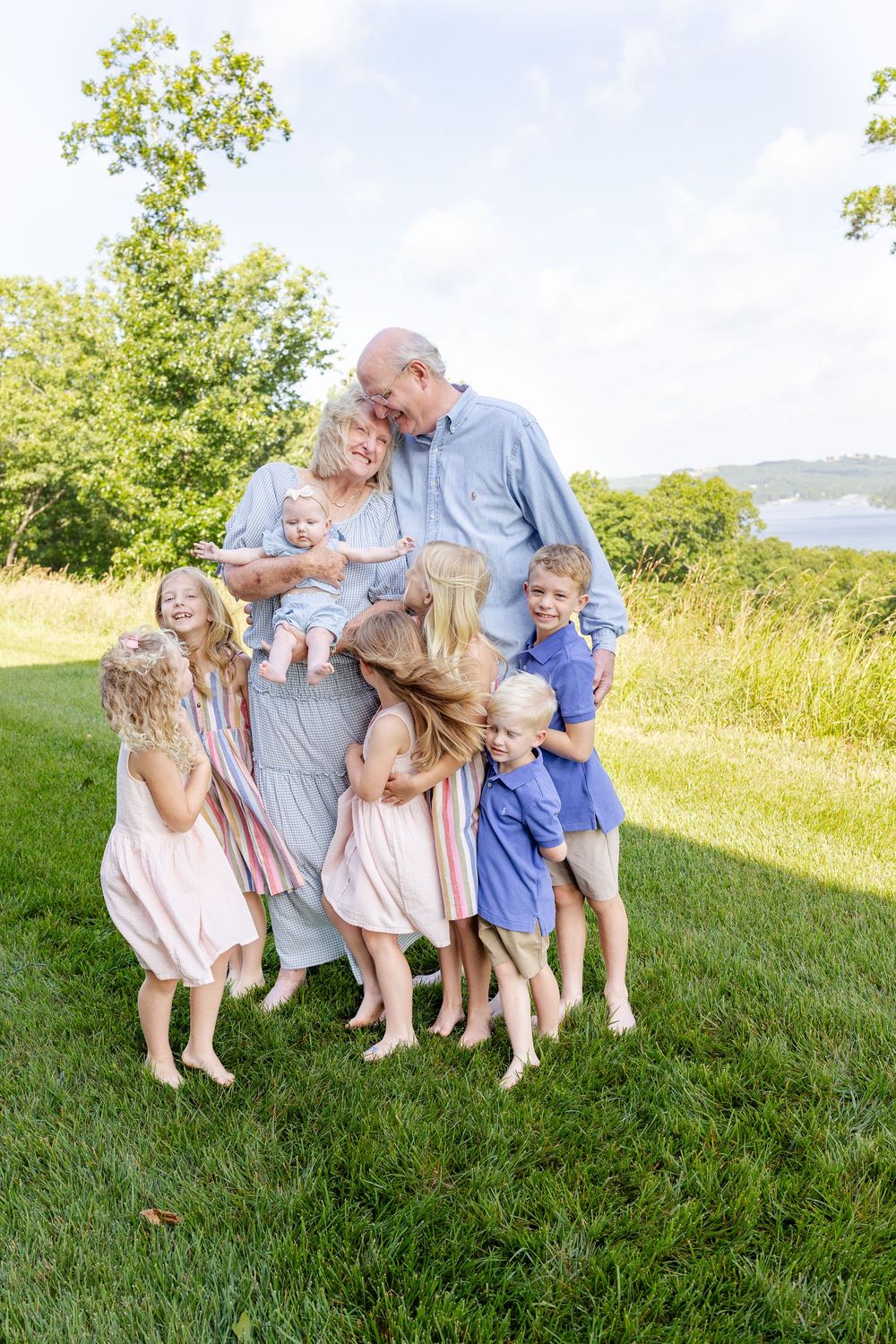 Grandparents surrounded by seven grandchildren hugging them on a green lawn with trees in the background.