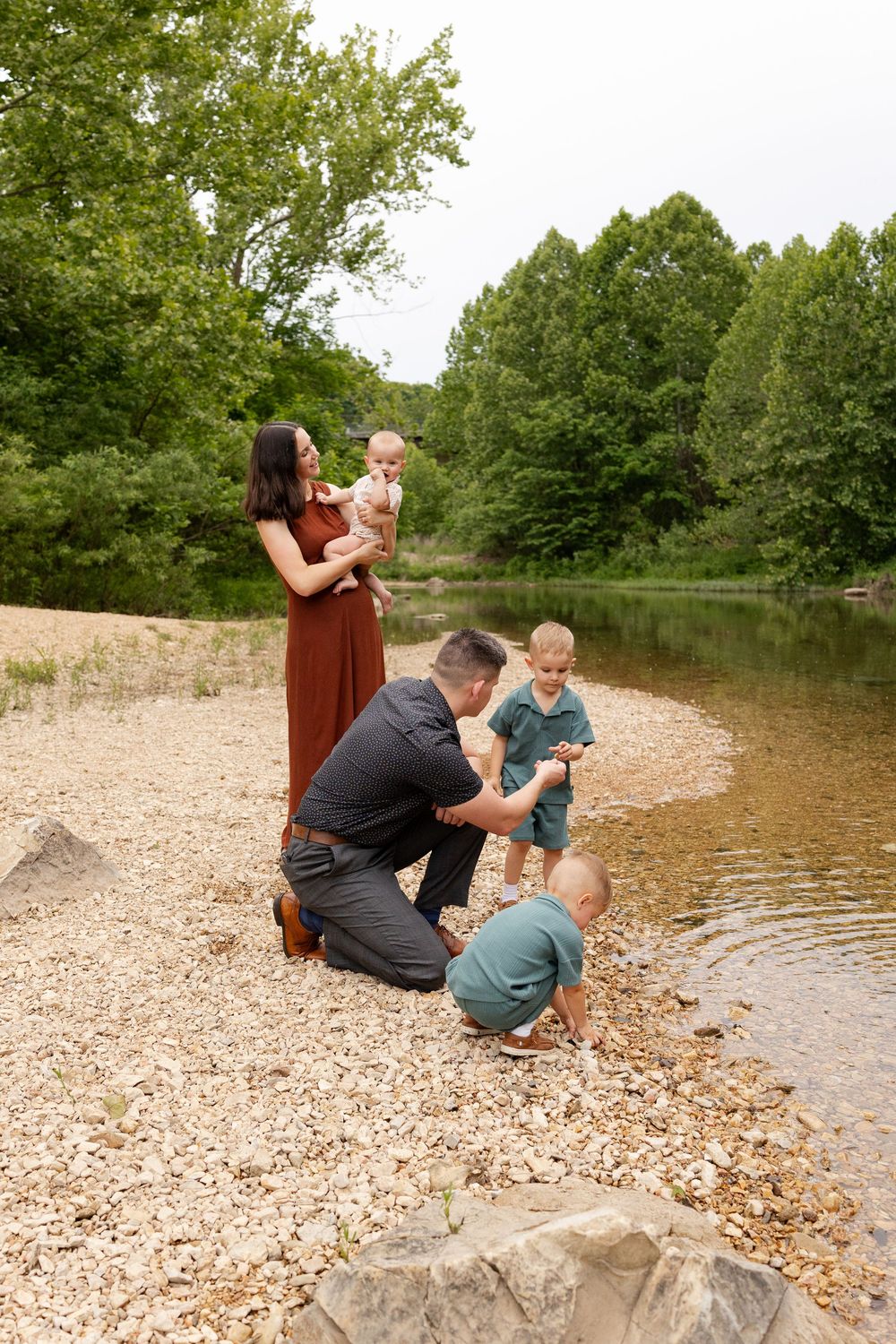 Family of five enjoying a peaceful moment by a rocky riverbank surrounded by lush green trees.
