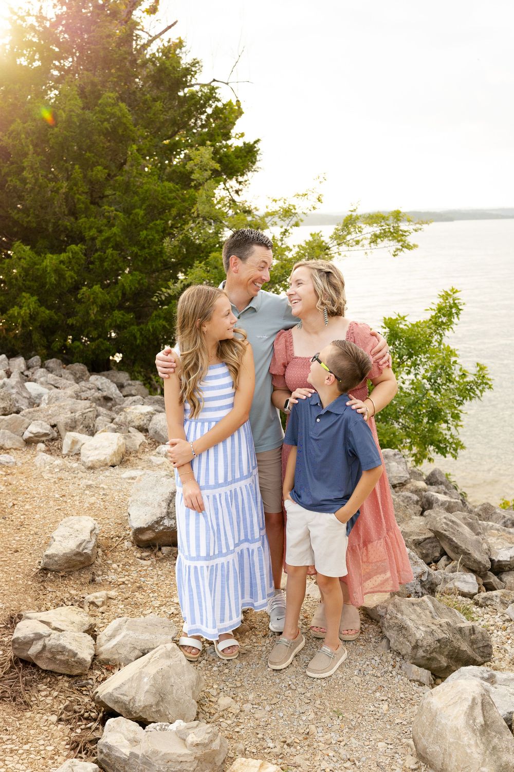 Family of four smiling together on rocky lakeside path with lush trees and sunlit water in background.