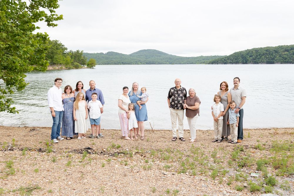 Large family group portrait on a sandy lakeside beach with green mountains and calm water in the background.