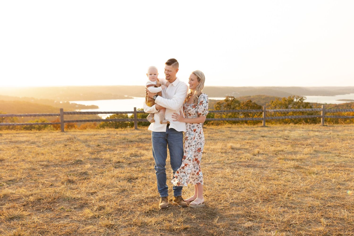 A happy family of three poses at golden hour on a grassy hill with a scenic water view in the background.