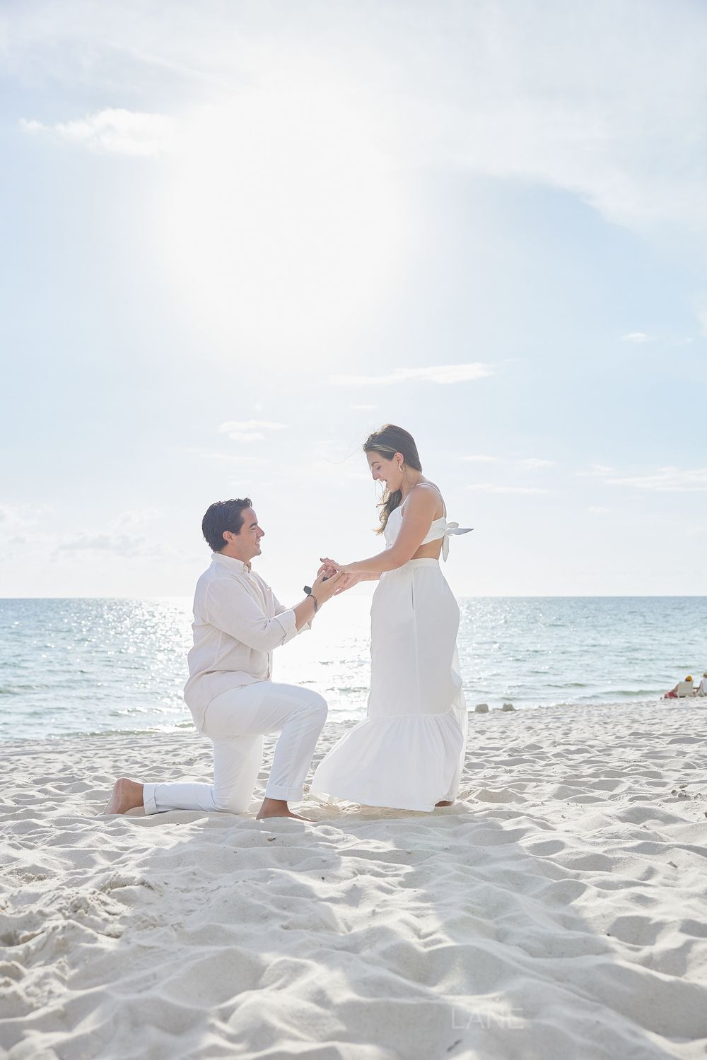 Man on one knee proposing to woman in white dress on a sunny sandy beach.