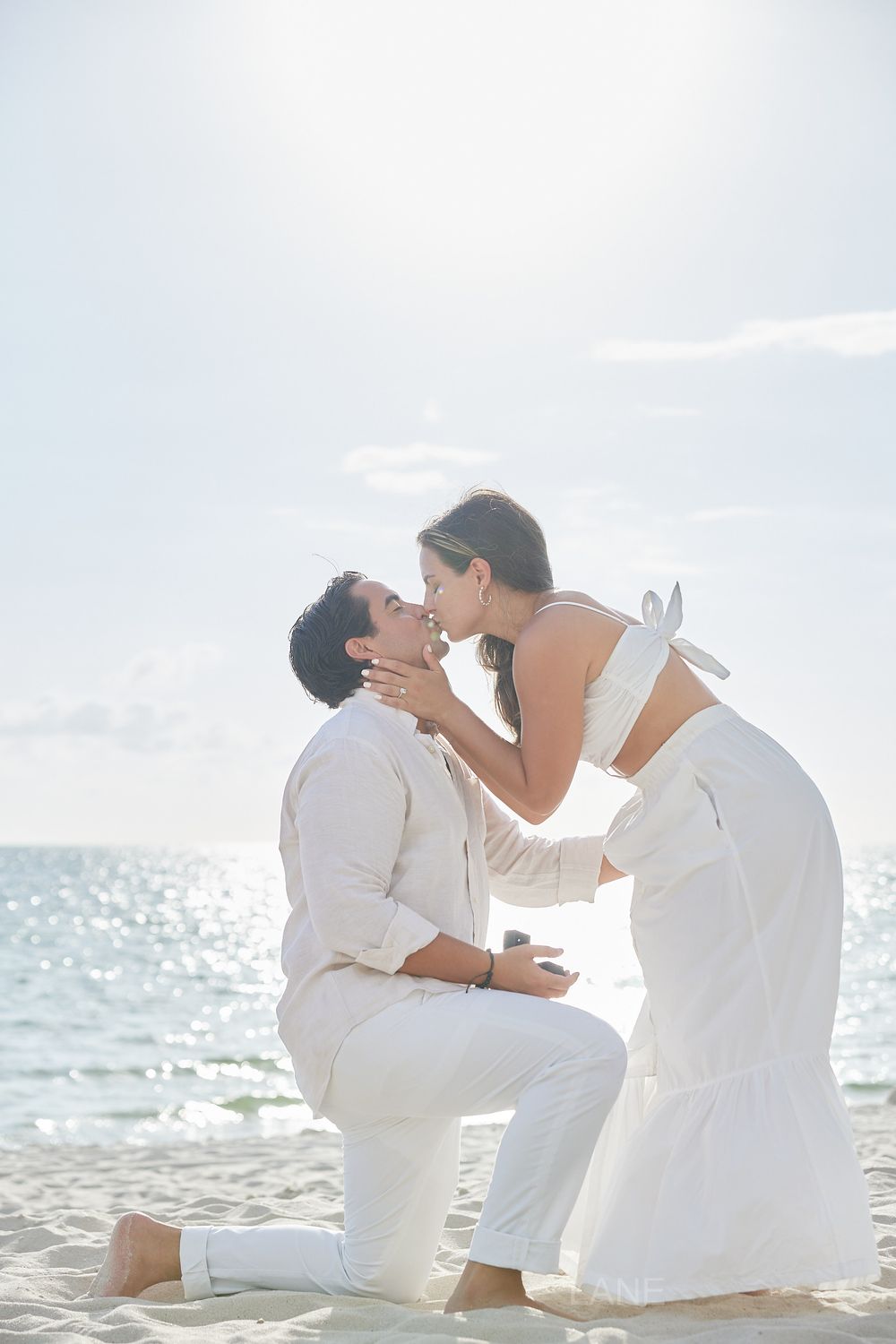 Couple sharing a romantic kiss on a bright sunny beach during a marriage proposal.