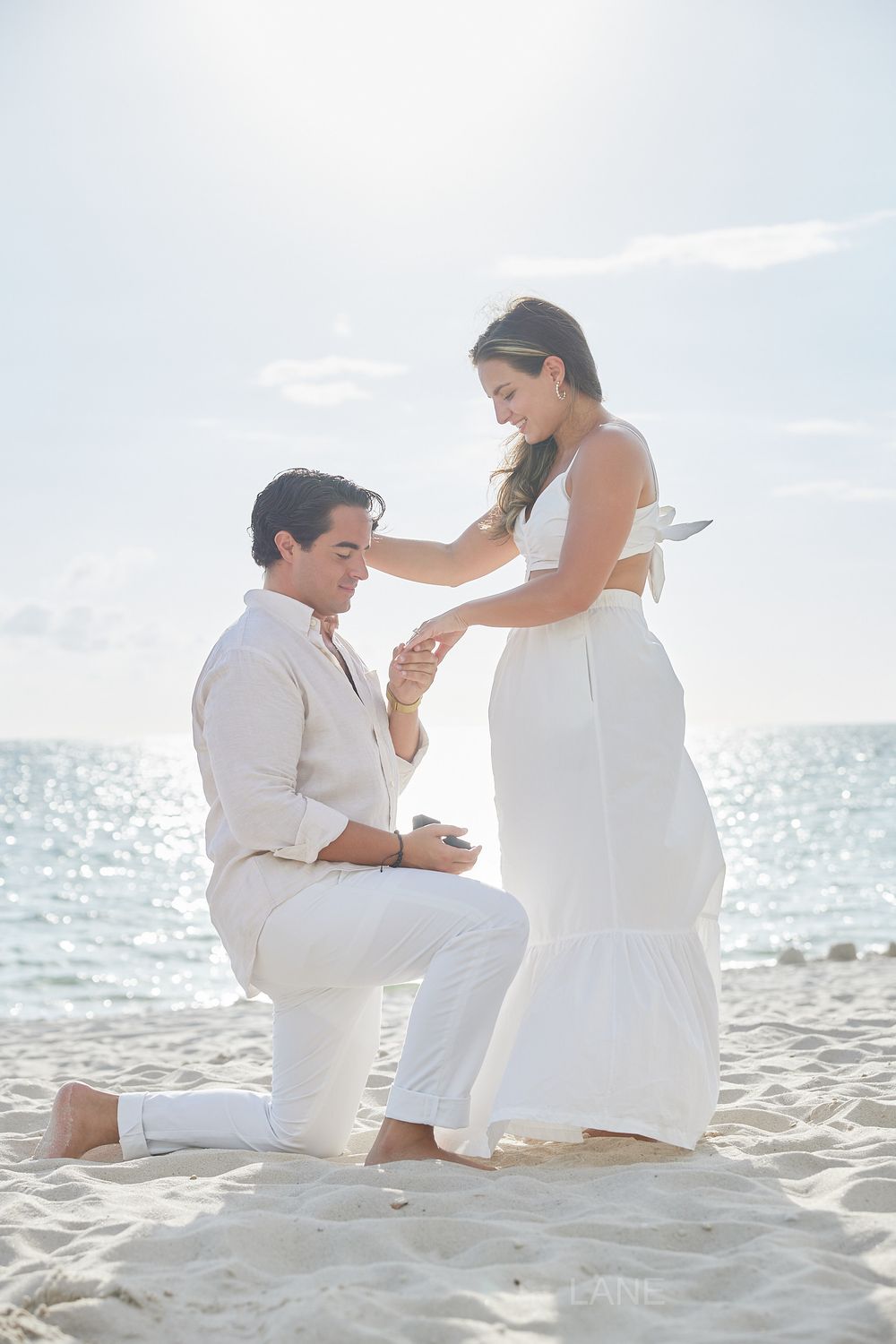 Man placing engagement ring on woman's finger while kneeling on a sandy beach.