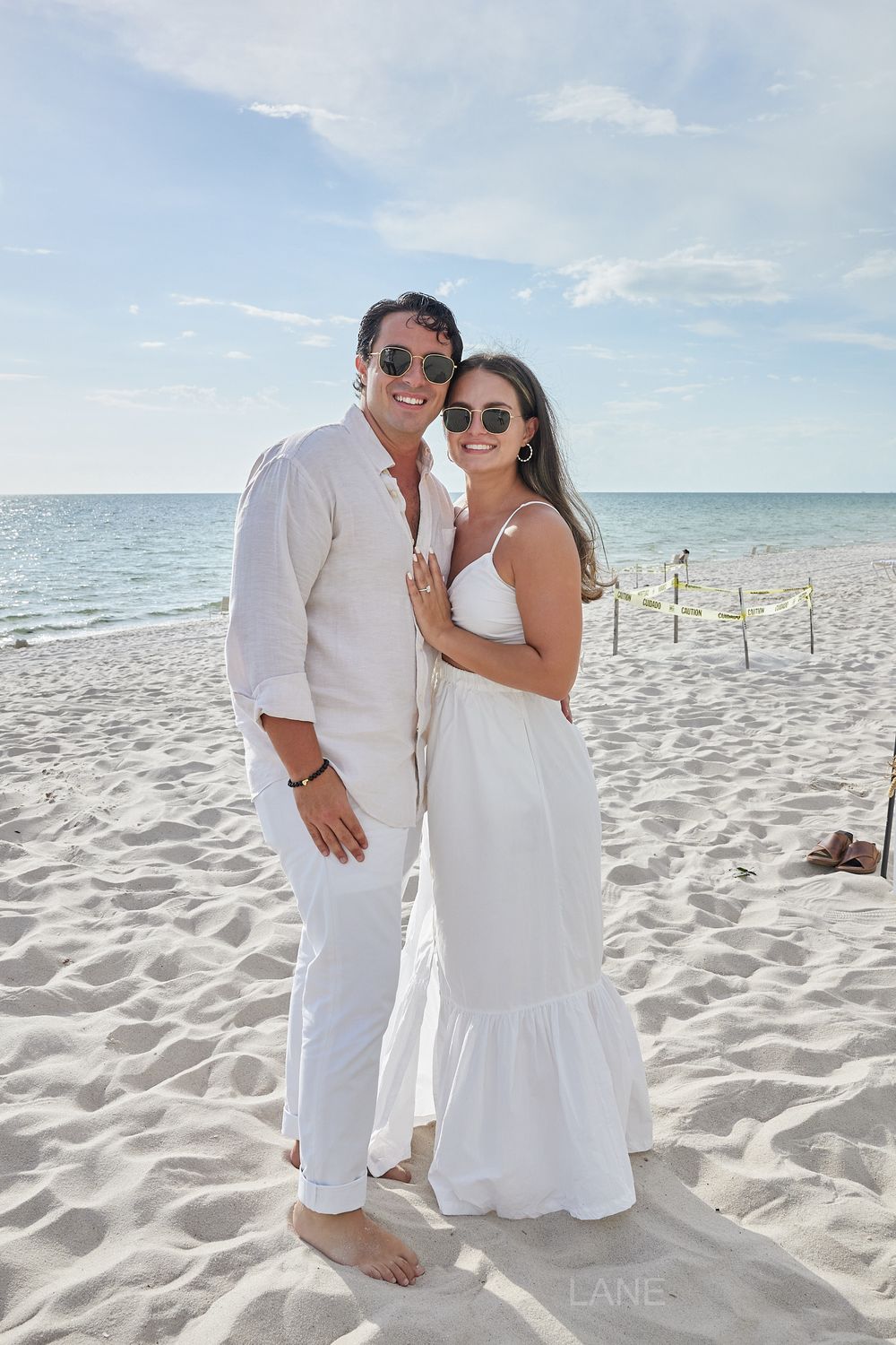 Smiling couple in matching white outfits posing together on a bright sandy beach.